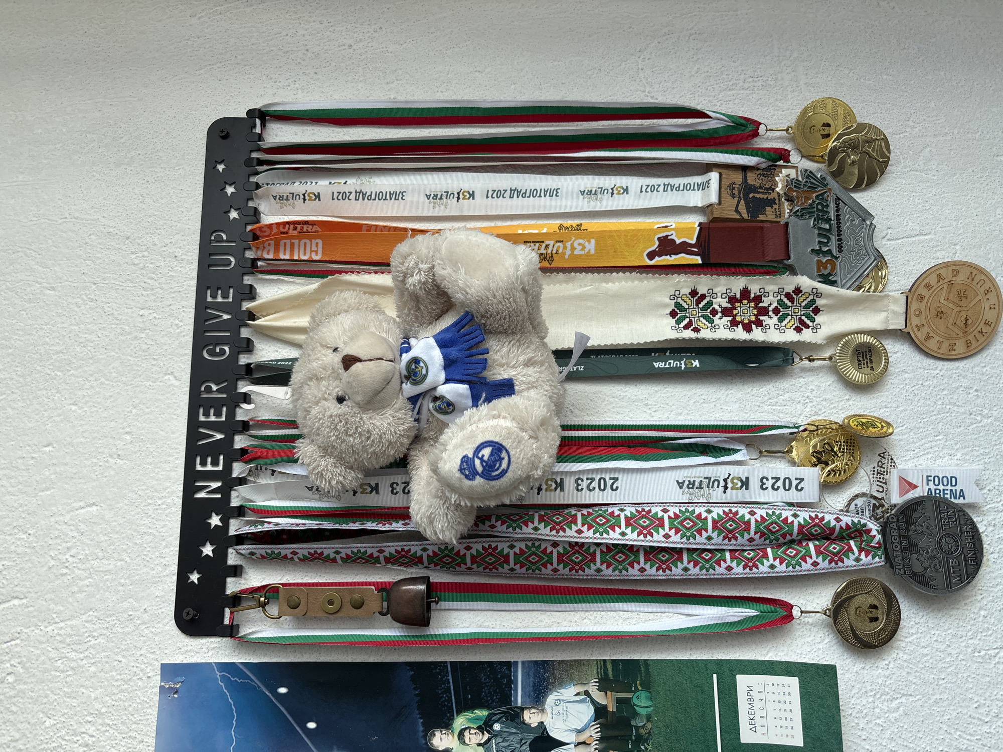 This image displays a black metal medal hanger labeled "NEVER GIVE UP," adorned with various medals and ribbons. Many ribbons are predominantly white with red and green stripes, reminiscent of the Bulgarian flag. Other ribbons have the text "ZLATOGRAD 2021 K3ULTRA" printed on them, indicating participation in the K3 Ultra race in Zlatograd, Bulgaria. There is also a medal or ribbon referencing "FOOD ARENA." A small, stuffed teddy bear wearing a blue and white scarf with a logo is nestled among the ribbons. Below the medal hanger, there are two items: a photograph of three young men in soccer attire and a calendar page with a month in Cyrillic script, presumably for December.