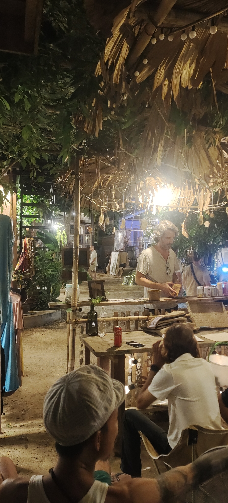 This media file depicts an outdoor evening scene at a casual establishment, likely a bar or restaurant, on Ko Pha-ngan, Thailand. A man with shoulder-length brown hair and a beard, wearing a light-colored v-neck t-shirt and glasses, is standing behind a wooden counter and appears to be engaged in preparing something, possibly food or a drink, as he holds a small wooden box. In front of him, on the counter, are several cans, some of which appear to be beverages, and a small potted plant. The setting is rustic and natural, with a thatched roof overhead adorned with string lights, and lush greenery surrounding the area.
In the foreground, the backs of two people are visible. One is wearing a checkered flat cap and a white tank top, with a beaded necklace. The other person is wearing a white t-shirt and dark pants, with visible tattoos on their arm. Several wooden tables and chairs are scattered around the sandy ground. A red Coca-Cola can and a smartphone are on one of the tables. The overall atmosphere appears relaxed and informal, suggesting a pleasant evening gathering. The lighting suggests it is nighttime, and the presence of dense foliage implies a tropical climate. The scene evokes a laid-back, possibly bohemian vibe, consistent with locations often found on Thai islands.