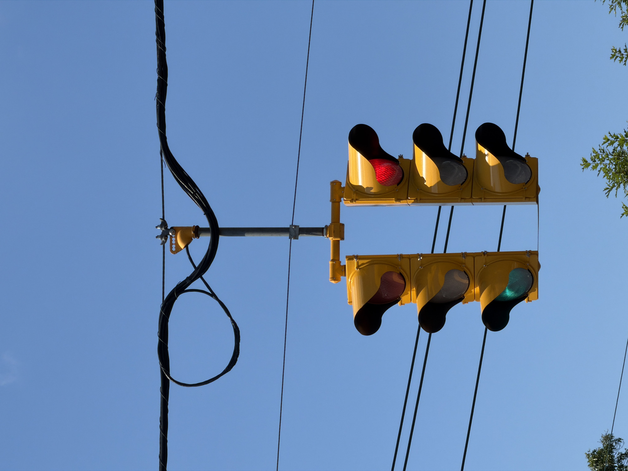 The image shows a traffic light signal suspended by wires against a clear blue sky. The traffic light has two vertical stacks of three lights each. The left stack has a red light illuminated at the top and a green light illuminated at the bottom. The right stack has a red light illuminated at the top and the middle and bottom lights are off. There are also power lines running horizontally across the frame. In the background, there is some greenery visible at the bottom of the image, suggesting it is outdoors. The scene is devoid of people or vehicles. The traffic light is a standard yellow, metal signal box. The wires appear to be electrical or communication cables. The overall environment appears to be urban or suburban, likely in Riverhead, United States of America, given the context.