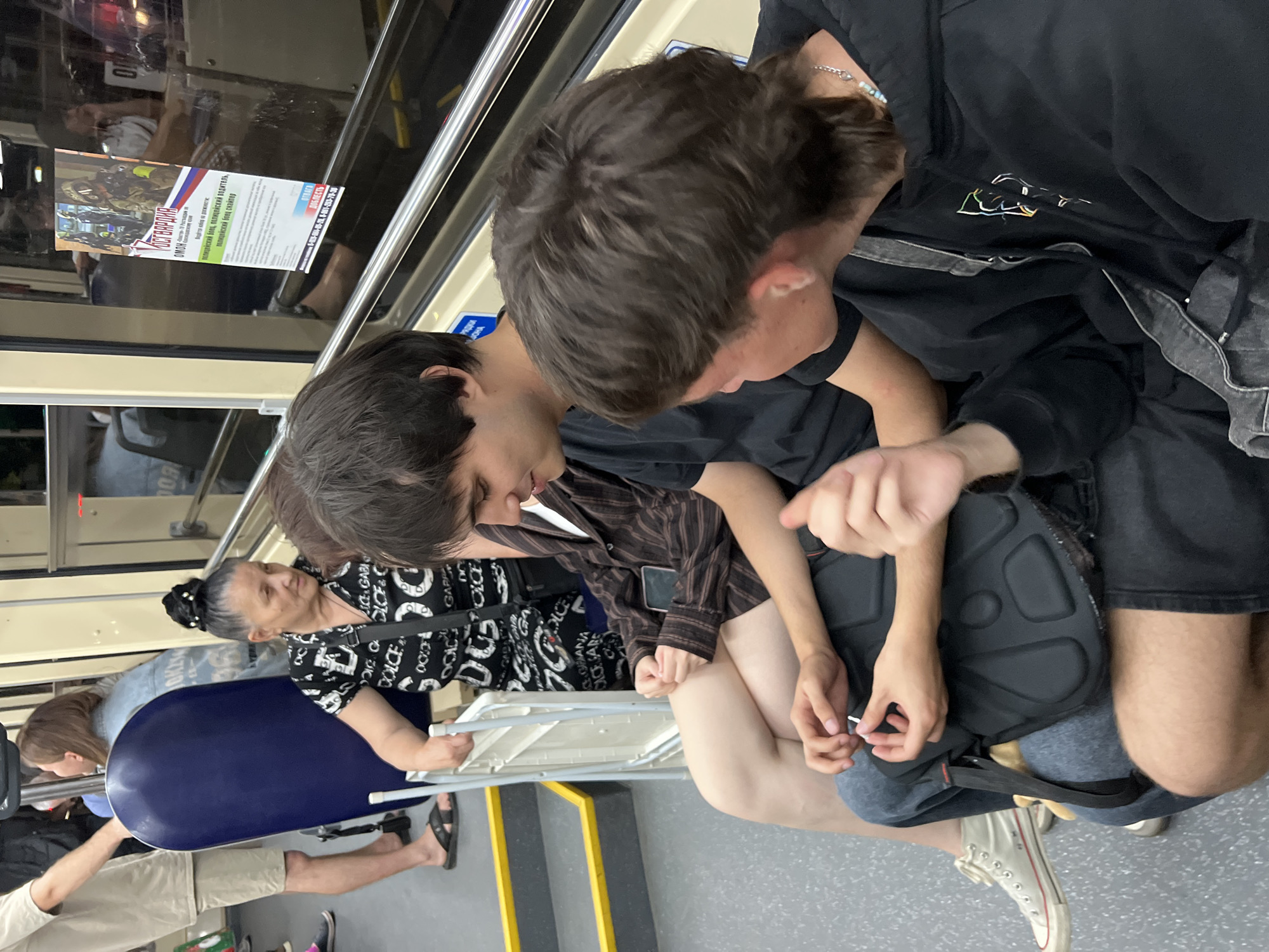 A group of people are seated inside a public transport vehicle, likely a bus or metro car, based on the interior design and the advertisement poster visible on the window. The advertisement, written in Russian, appears to be for recruitment into a law enforcement or security agency, featuring images of armed personnel.
In the foreground, two young men are seated. The one on the right has dark, mid-length hair styled in a mullet and wears a black hooded sweatshirt with a colorful graphic print and a silver chain necklace. The man on the left has darker, shorter hair and is wearing a dark t-shirt. Their hands are visible, suggesting they are engaged in conversation or interaction.
Behind them, an older woman with her hair pulled up in a bun wears a black and white patterned dress. Her arms are resting on a white folding seat. She is looking to her right. To her left, another individual, a young woman with fair skin, is seated, wearing a brown and black striped shirt and jeans. Her legs are crossed, and a phone is visible on her lap.
Further in the background, the arm and upper body of a person wearing a beige shirt and shorts are visible, as are parts of other passengers and the vehicle's interior, including yellow and grey flooring and metal railings. The scene appears to be taking place during daylight hours. The presence of a specific advertisement for Russian law enforcement suggests the location is within Russia, and the context of public transport in a Russian city like Krasnodar is plausible.