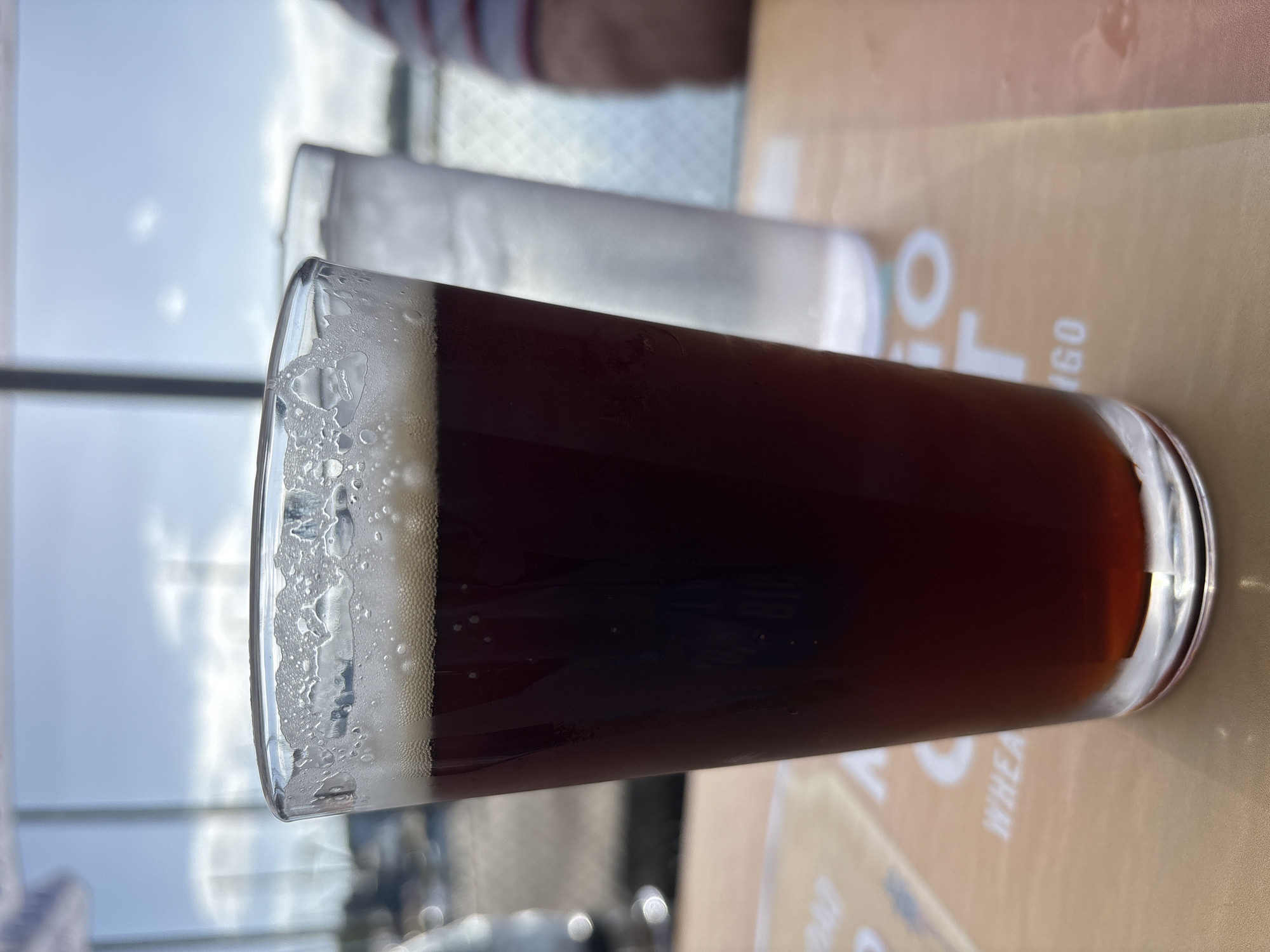 A close-up shot focuses on a pint of dark brown beer in a glass with a frothy head, set on a table. Behind it, a second glass of water is visible, along with a blurred background of a sunny outdoor scene with a body of water and some structures. The table is covered with a placemat or menu with white lettering that is partially obscured. The lighting suggests it is daytime, likely late morning or afternoon.