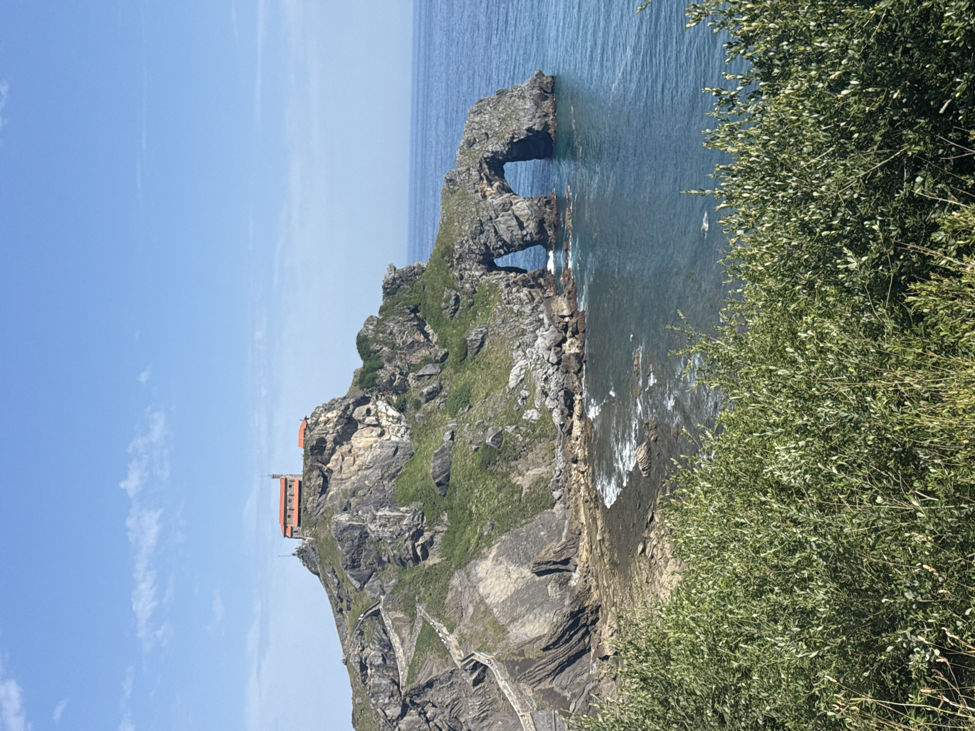 The image depicts the island of San Juan de Gaztelugatxe, a unique coastal landmark located off the coast of Bermeo, Spain. A prominent feature is the hermitage of San Juan, situated atop the rocky island, identifiable by its distinctive red-tiled roof.
Access to the island is provided by a long, winding stone staircase that ascends the steep cliffs. The staircase is a visible element in the scene, snaking its way up the rugged terrain.
Adjacent to the island, a natural rock formation creates a double archway, allowing the sea to pass through. This archway is a dramatic geological feature contributing to the dramatic seascape.
The island is surrounded by the deep blue waters of the ocean, with gentle waves lapping against the rocky shore. The sky above is a clear, bright blue with a few wispy white clouds.
The foreground of the image is dominated by lush green foliage, likely bushes and trees, suggesting a natural, undeveloped environment. The overall scene is a picturesque representation of the natural beauty and architectural significance of San Juan de Gaztelugatxe.