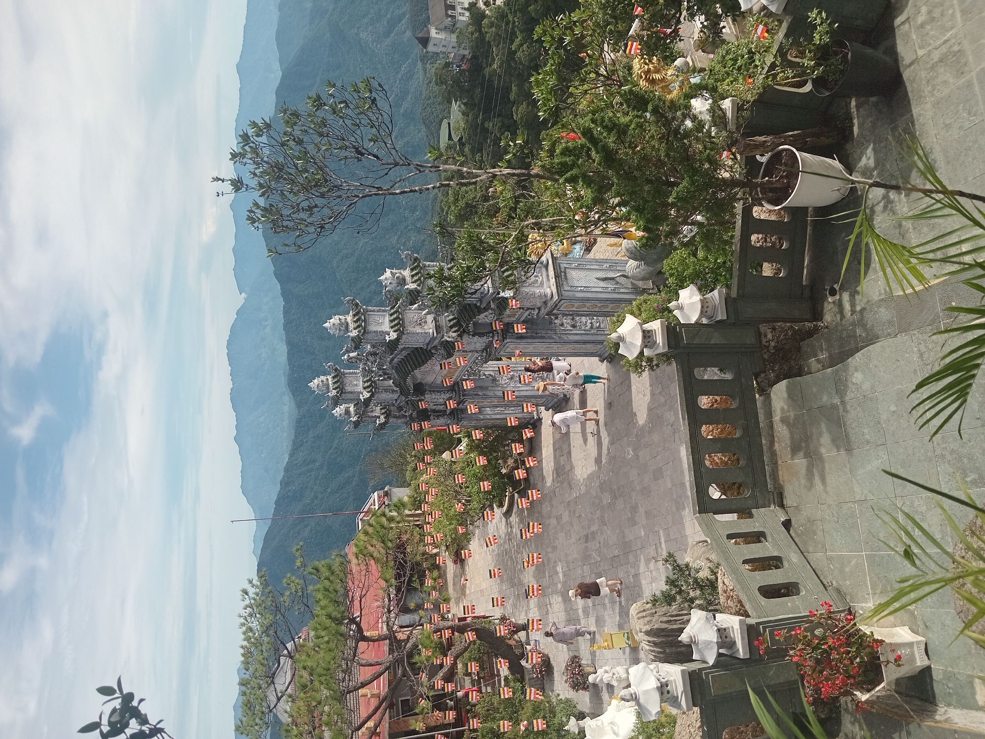 This image shows a view from a higher elevation overlooking a temple complex and a mountainous landscape. In the foreground, there is a gray stone railing with decorative cutouts, with potted plants on either side. To the left, a white pot contains bright red flowers. Steps lead down from the foreground.
Further into the scene, a paved courtyard is visible, adorned with numerous small flags strung between trees and poles. The flags are rectangular and feature horizontal stripes of yellow, red, and white. A large, ornate gray gate with multiple tiers and decorative architectural elements stands prominently in the mid-ground. People, dressed in casual clothing, are walking in the courtyard. To the left of the gate, a building with a red tiled roof is partially visible.
The background reveals a lush, mountainous landscape with rolling hills covered in dense green vegetation. The sky above is partly cloudy with patches of blue. In the distance, several layers of mountains recede into the haze, creating a sense of depth. The architecture of the temple and the surrounding environment suggest a location in a mountainous or hilly region, likely a religious or cultural site. The presence of flags and traditional architectural elements could indicate a Southeast Asian setting.
This scene is consistent with the Ba Na Hills area near Đà Nẵng, Vietnam, which is known for its European-inspired architecture, Buddhist temples, and stunning mountain vistas.