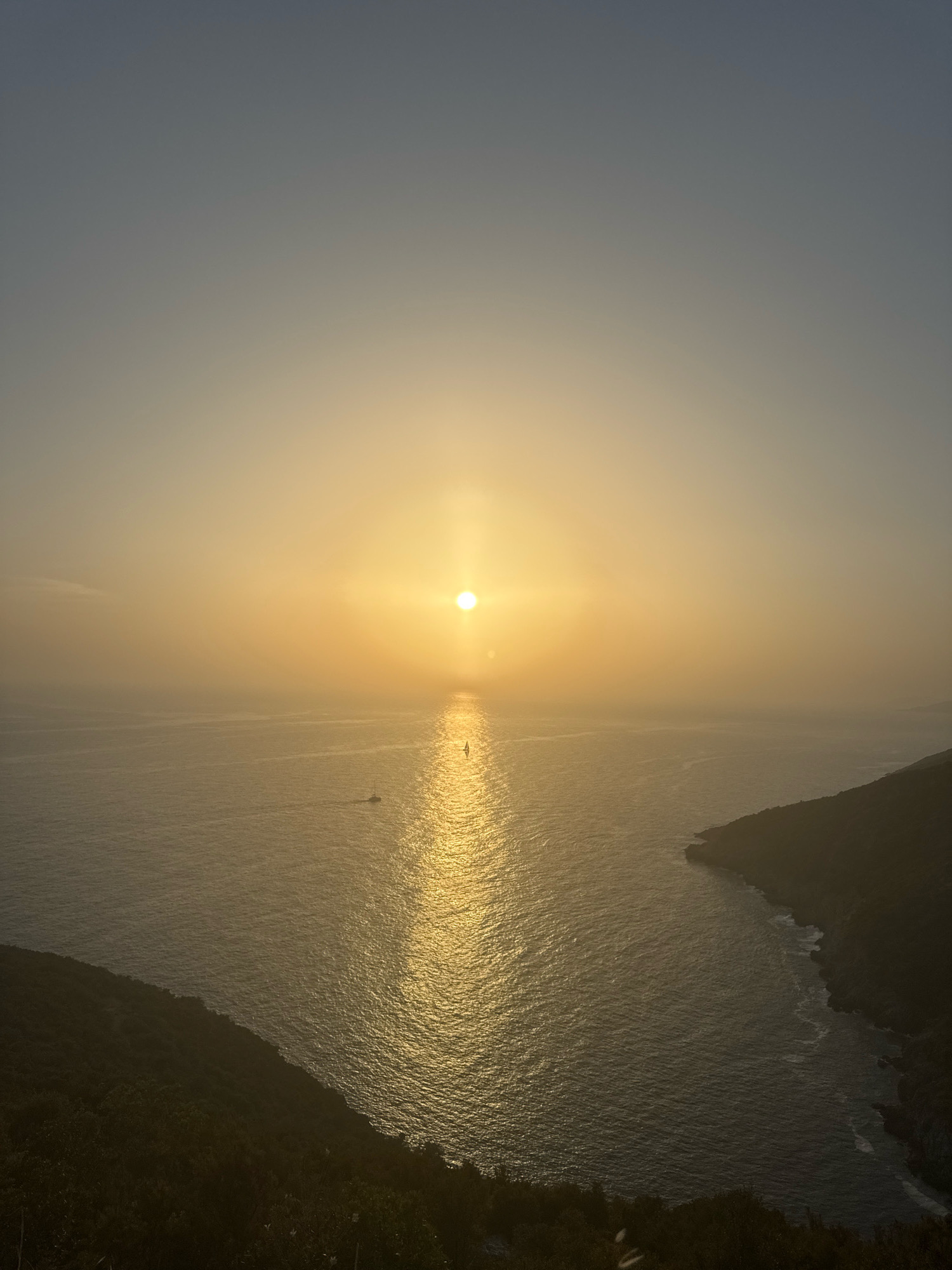 An aerial, high-angle shot captures a serene sunset over the Adriatic Sea near Zagora, Montenegro. The sun, a bright, glowing orb, is positioned slightly left of the frame's center, casting a shimmering golden path across the tranquil water. A small sailboat is visible on the sea, its silhouette distinct against the radiant reflection of the sun. Lush, dark green hills flank the water, with one prominent headland curving into the sea on the right side of the image. The sky transitions from a pale yellow near the horizon to a soft grey at the top, with a slight haze obscuring the distant coastline. The overall atmosphere is one of peaceful natural beauty.