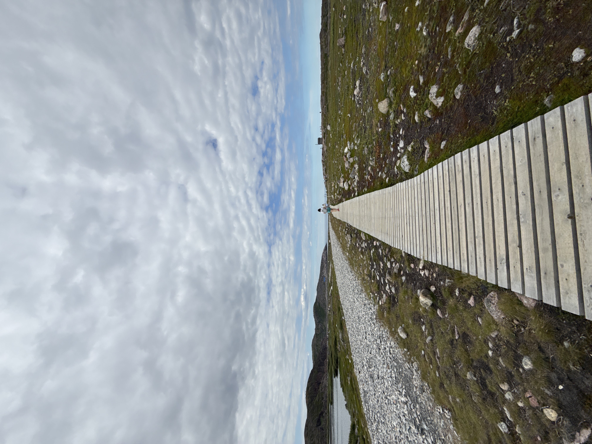 A person walks on a wooden boardwalk that extends into the distance. The boardwalk is bordered by a rocky, grey terrain on the left and a green, mossy landscape dotted with rocks on the right. In the background, a body of water is visible to the left, and hills can be seen beyond it. The sky is cloudy with patches of blue sky. There are a few small structures visible in the far distance. This scene depicts a path in a natural, possibly coastal, environment.