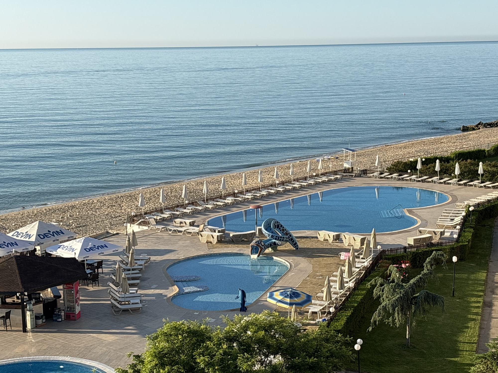 This is an elevated, wide shot of a resort area on the coast of Varna, Bulgaria. The image features a calm, blue sea stretching to the horizon under a clear, pale blue sky. The beach is a strip of light brown sand, lined with rows of white beach umbrellas and lounge chairs. Beyond the beach, there are two interconnected swimming pools. The larger pool is a vibrant blue, and the smaller one has a distinct kidney shape with a shallow section. A playful blue water slide, shaped like a serpent or dragon, is situated in the smaller pool, and a dolphin statue is also present. Around the pools, there are more lounge chairs and umbrellas, as well as a bar area with a dark roof and a red refrigerator. Lush green trees and manicured hedges border the resort area. The overall scene depicts a peaceful, sunny day at a seaside resort.