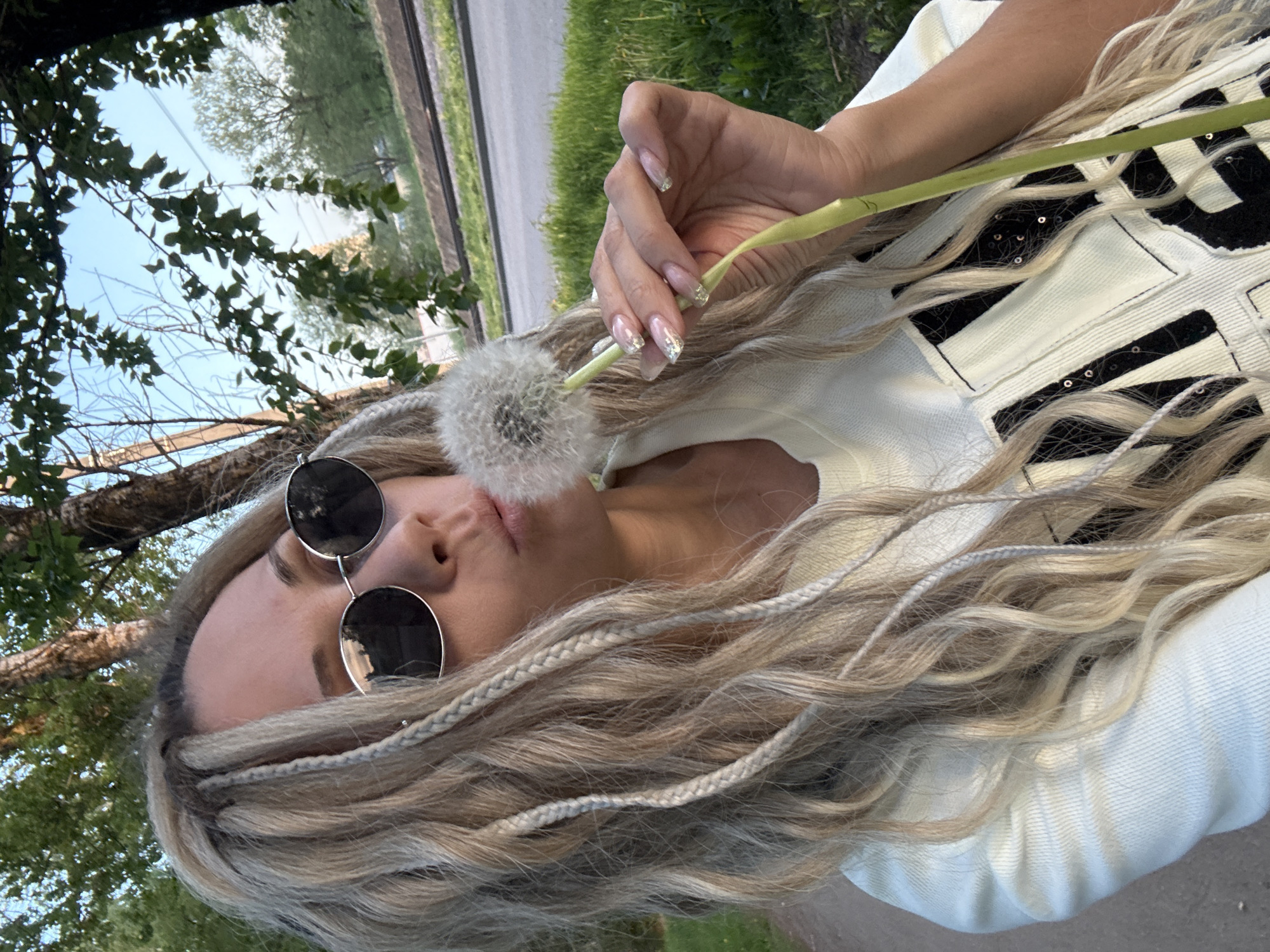 A woman with light blonde braided hair and round sunglasses is holding a dandelion in front of her face. She has manicured nails with a sparkly design. She is wearing a white top with black embellishments. The background shows a street, grass, and trees, suggesting an outdoor location. The city is Rusanovo, Russia.