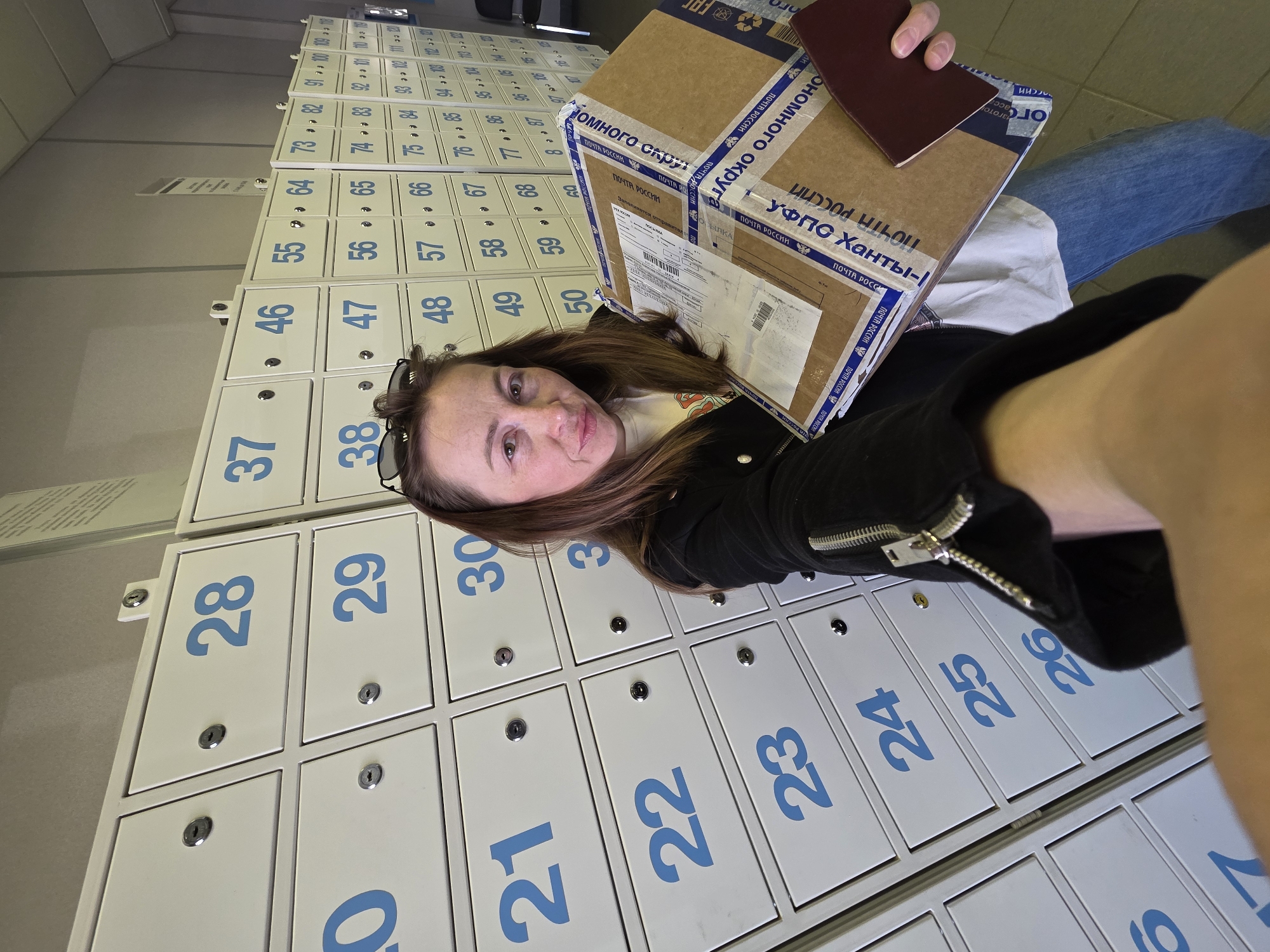 A female individual is depicted taking a selfie inside a Russian Post office in Chaikovsky, Russia. She is approximately in her 30s to 40s, with light brown hair and dark sunglasses resting atop her head. She wears a black jacket with a silver zippered sleeve, a white t-shirt, and blue jeans, displaying a slight smile. Her right arm is extended, holding the camera. In her left arm, she holds a brown cardboard package secured with blue and white "Почта России" (Russian Post) branded adhesive tape and visible shipping labels. Concurrently, she holds a dark red passport or similar document in her left hand. The background features a wall composed of multiple vertical columns and horizontal rows of white, locked post office boxes, each individually numbered in blue, ranging from the low 20s on the left to at least 122 on the far right. This arrangement is characteristic of a postal service customer area for mail and parcel management.
