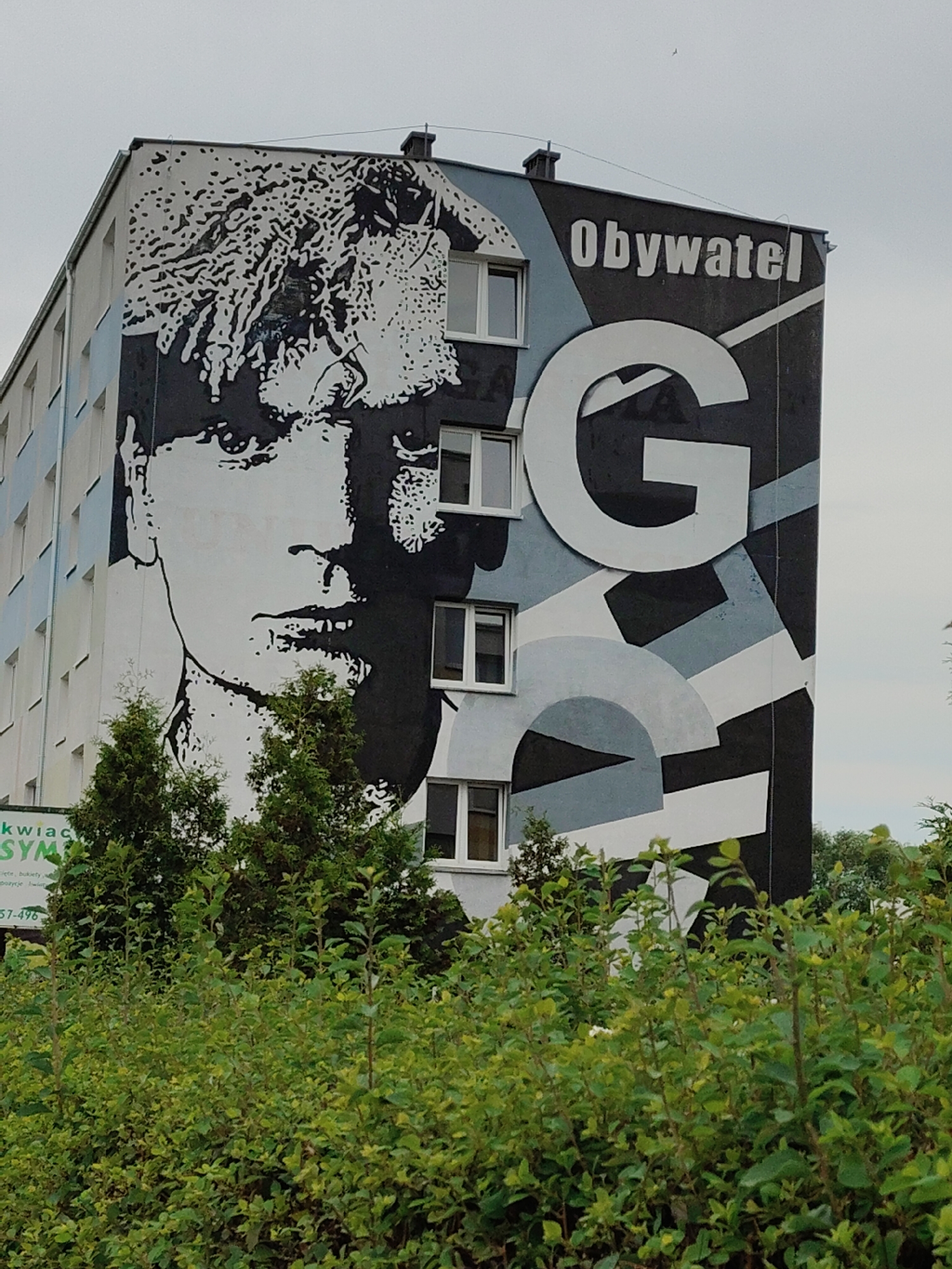 A large mural covers the side of a tall, multi-story building. The mural features a black and white portrait of a person's face, with stylized hair. To the right of the face is a large white letter "G" and the word "Obywatel" (Citizen in Polish). The building has multiple windows visible. In the foreground, there are lush green bushes and trees obscuring the lower portion of the building. The sky above is overcast. There is a sign partially visible on the left side of the building that says "kwaciak SYM" and a phone number "57-496".