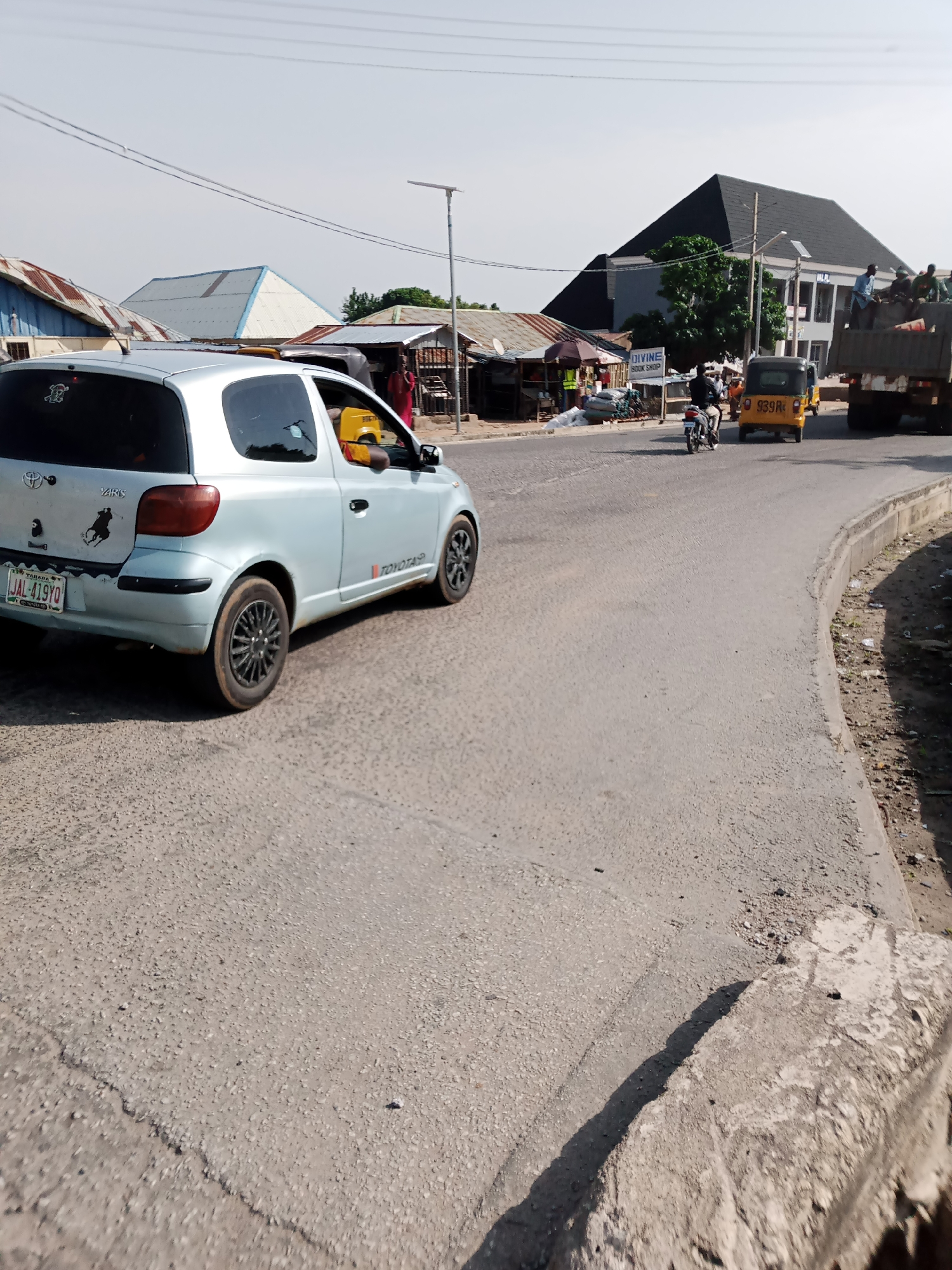 The image depicts a street scene in Jalingo, Nigeria, on a clear day. In the foreground, a light blue Toyota Yaris car is visible from the rear-left quarter, facing forward. The car has a license plate that reads "JAL-419YO". A person is partially visible in the driver's seat. To the right of the Yaris, a yellow auto-rickshaw (Keke Napep) with the license plate "939R" is traveling in the same direction. Further to the right, a large truck loaded with people and goods is moving away from the camera. The background features several buildings with corrugated metal roofs and some with concrete structures, suggesting a commercial or residential area. A "DIVINE BOOK SHOP" sign is visible on the left side of the road. The road surface appears to be asphalt with some wear and tear. Utility poles and overhead wires are present in the sky. The scene captures everyday street life with various modes of transportation and roadside establishments.