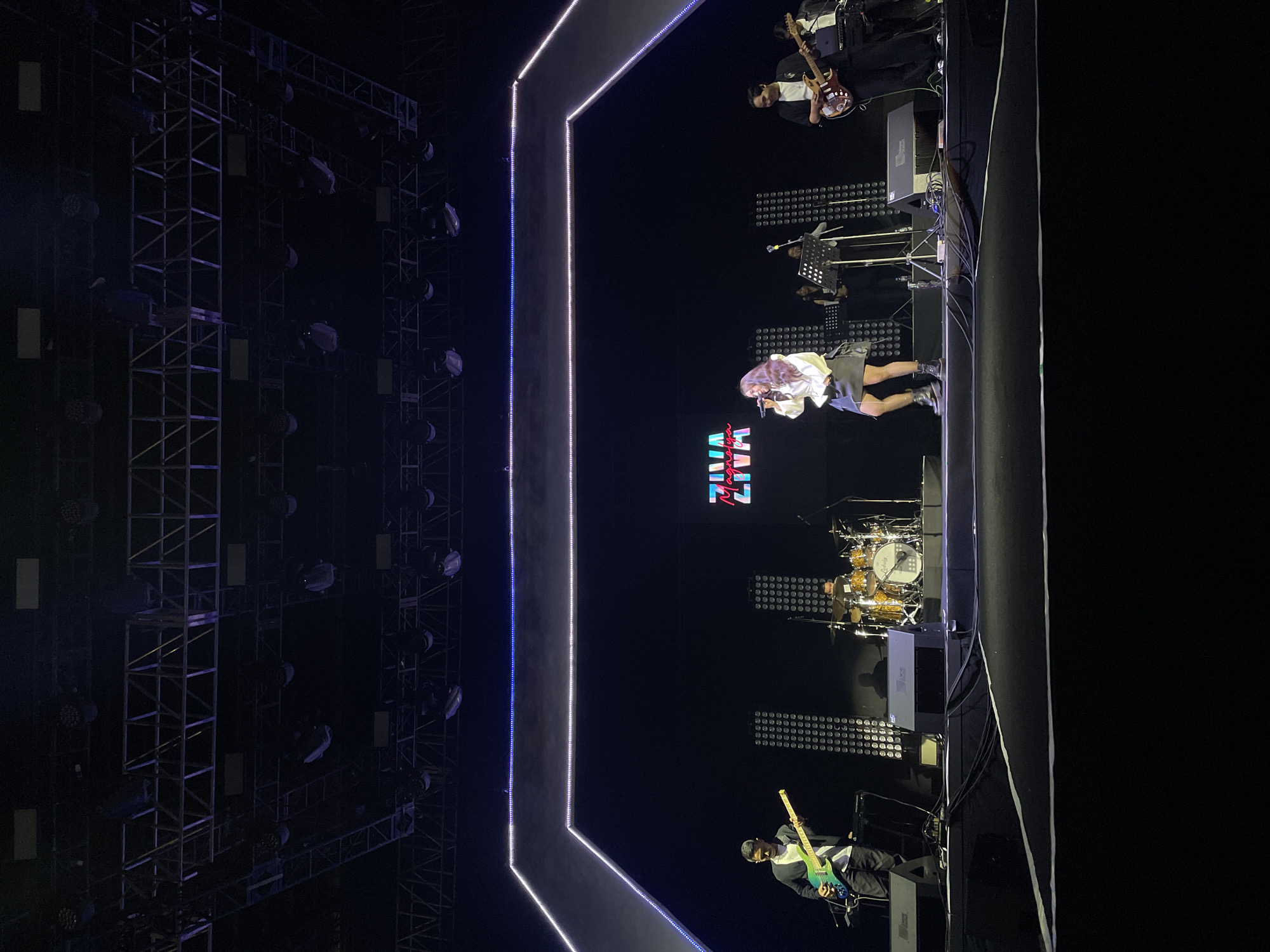 The image shows a musical performance at a venue with a stage in Salembaran, Indonesia. A female vocalist with pink hair is performing on stage, backed by a band consisting of two guitarists, a drummer, and other musicians. The stage is decorated with neon lights. The lighting is dark, with spotlights on the performers. A large scaffolding structure is on the left.