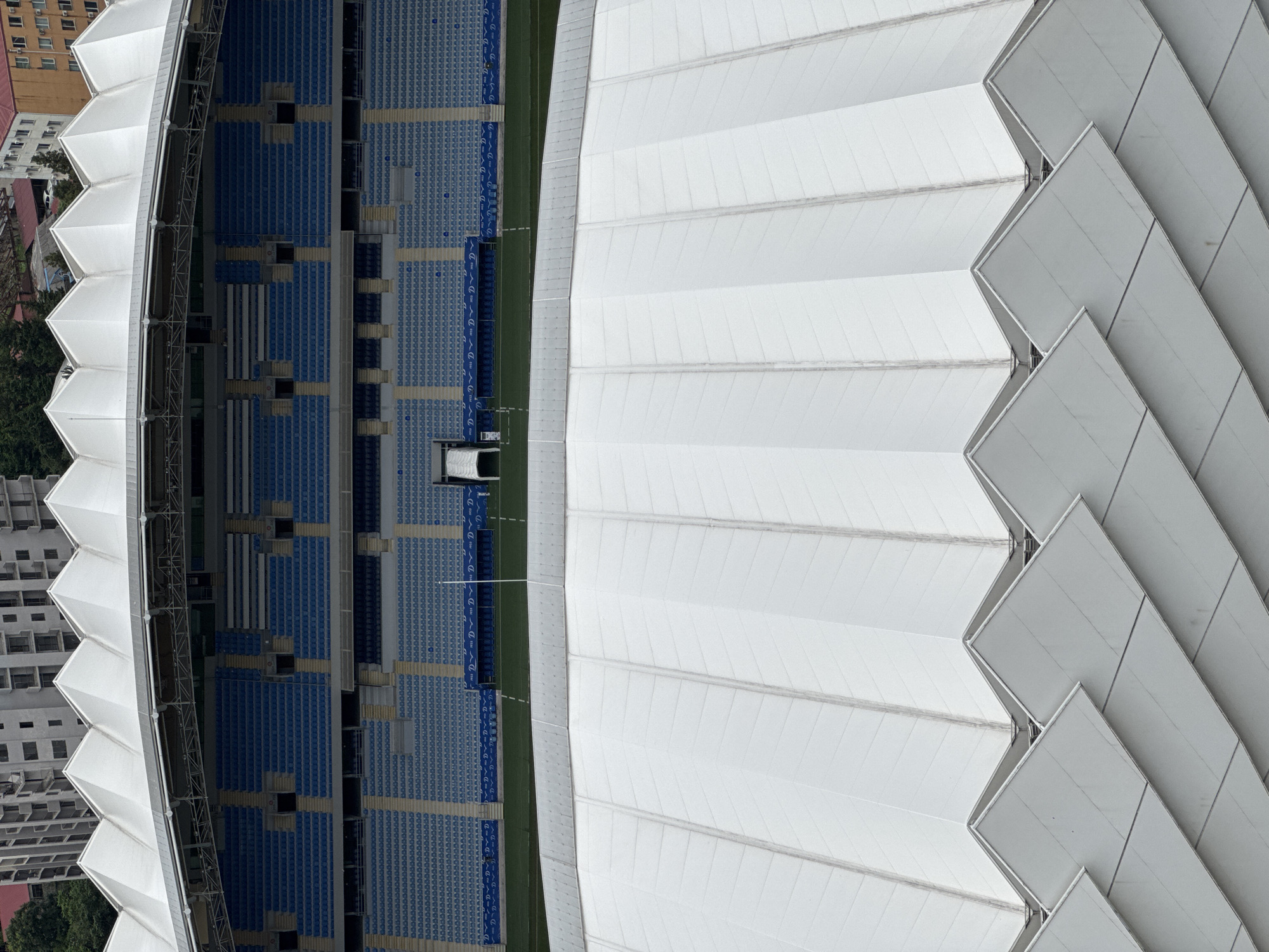 The image shows a high-angle, direct view of a modern stadium in Batumi, Georgia. The foreground is dominated by the white, angular, pleated roof of the stadium, designed with a repeating pattern of inverted V-shapes. Beyond the white roof, the tiered blue seating of the stadium is visible, interspersed with light-colored vertical supports and dark rectangular openings. The field of the stadium is a strip of green grass situated below the seating. In the background, several multi-story buildings with light-colored facades and dark windows are visible, suggesting an urban environment. The sky is overcast. There are no people or any discernible activity depicted in the scene.
