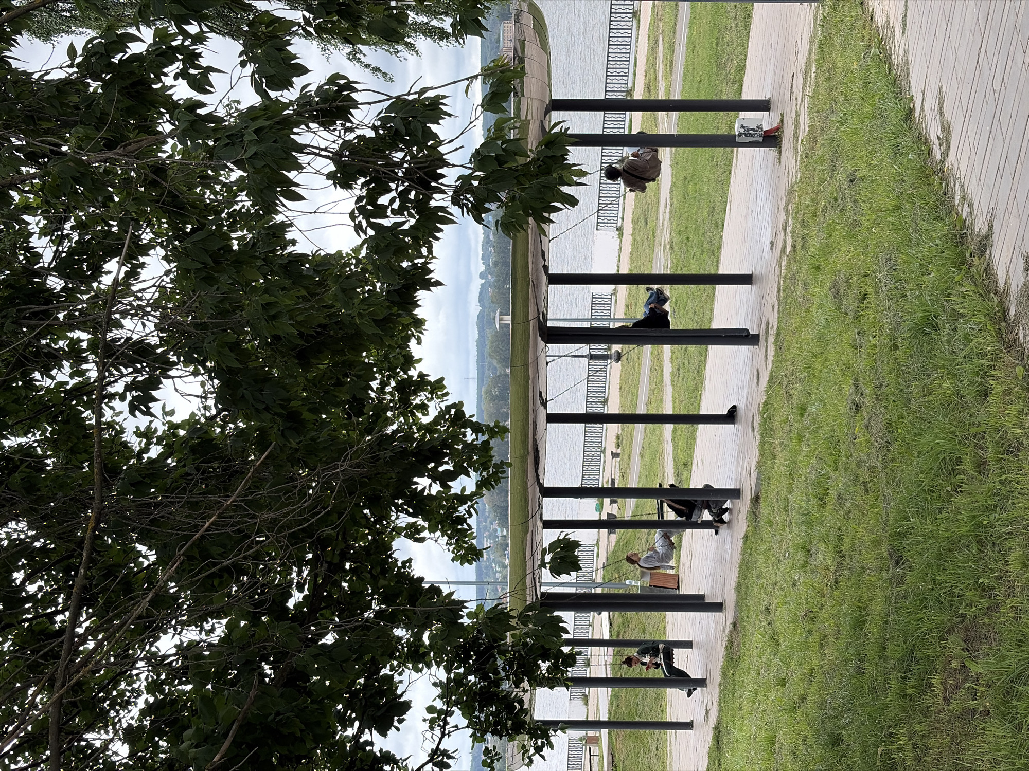 The image shows a park scene in Votkinsk, Russia, featuring a covered swing set area along a body of water, likely the Votkinsk Reservoir. Four individuals are visible on swings. In the foreground, a person sits on a swing, looking at a phone. To their right, another person on a swing is also looking down, possibly at a device. Further to the right, a third person is on a swing, leaning back with their eyes closed. A fourth person is on a swing in the far right of the frame, also leaning back. The swings are under a modern, curved roof supported by dark pillars. Beyond the swing area, a paved walkway runs alongside the water, which is bordered by a black metal fence. Green grass and trees fill the rest of the park. In the distance, across the water, the cityscape of Votkinsk can be seen, including buildings and a tall white tower. The sky is overcast.