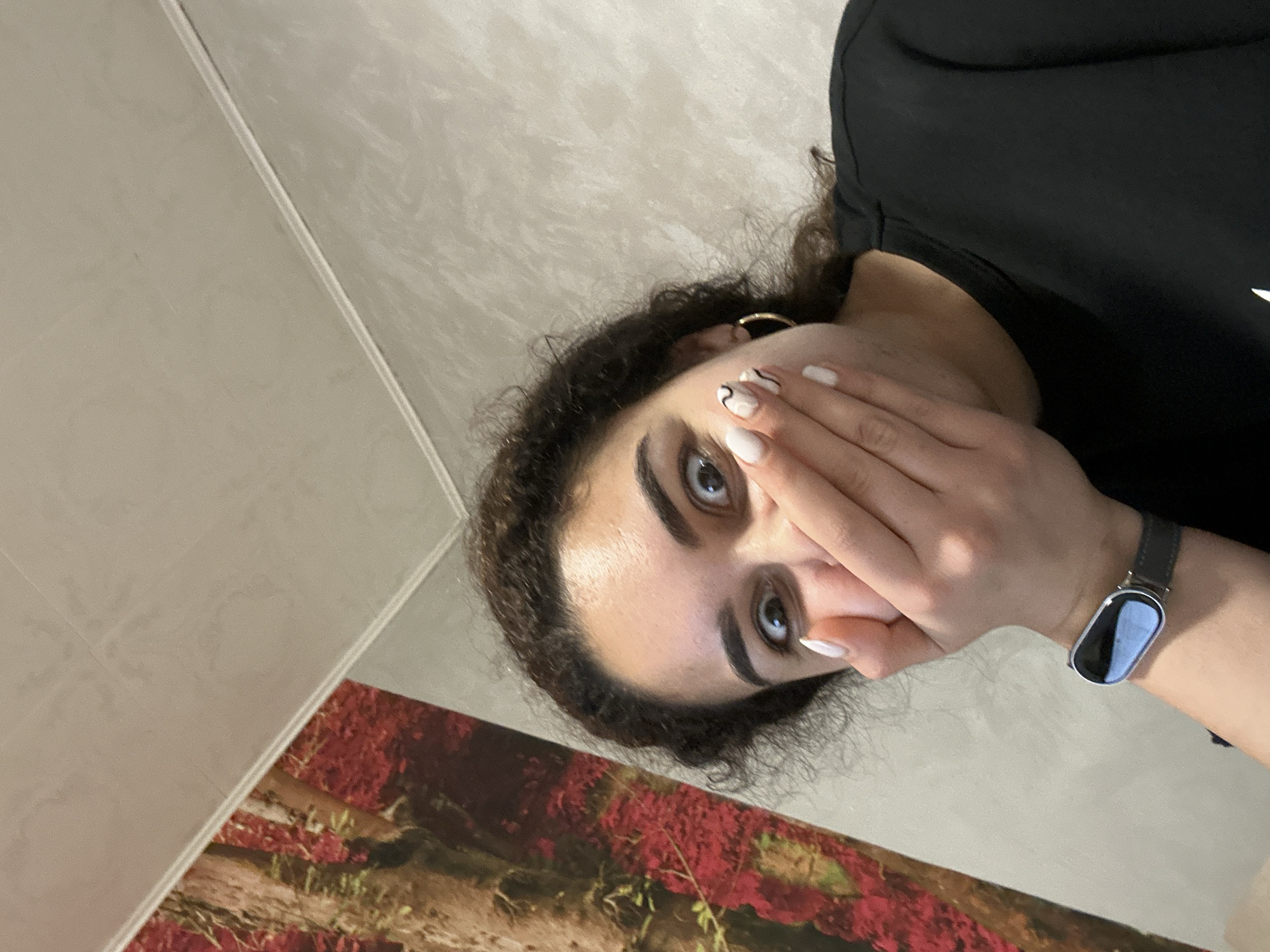 A woman with curly dark hair and fair skin covers part of her face with her left hand. She wears a black t-shirt and a silver smartwatch on her left wrist. The fingernails on her left hand are painted white with black squiggle designs on some. She appears to be indoors with a wall featuring a floral artwork behind her. There is no indication of the location being in Burgas, Bulgaria.