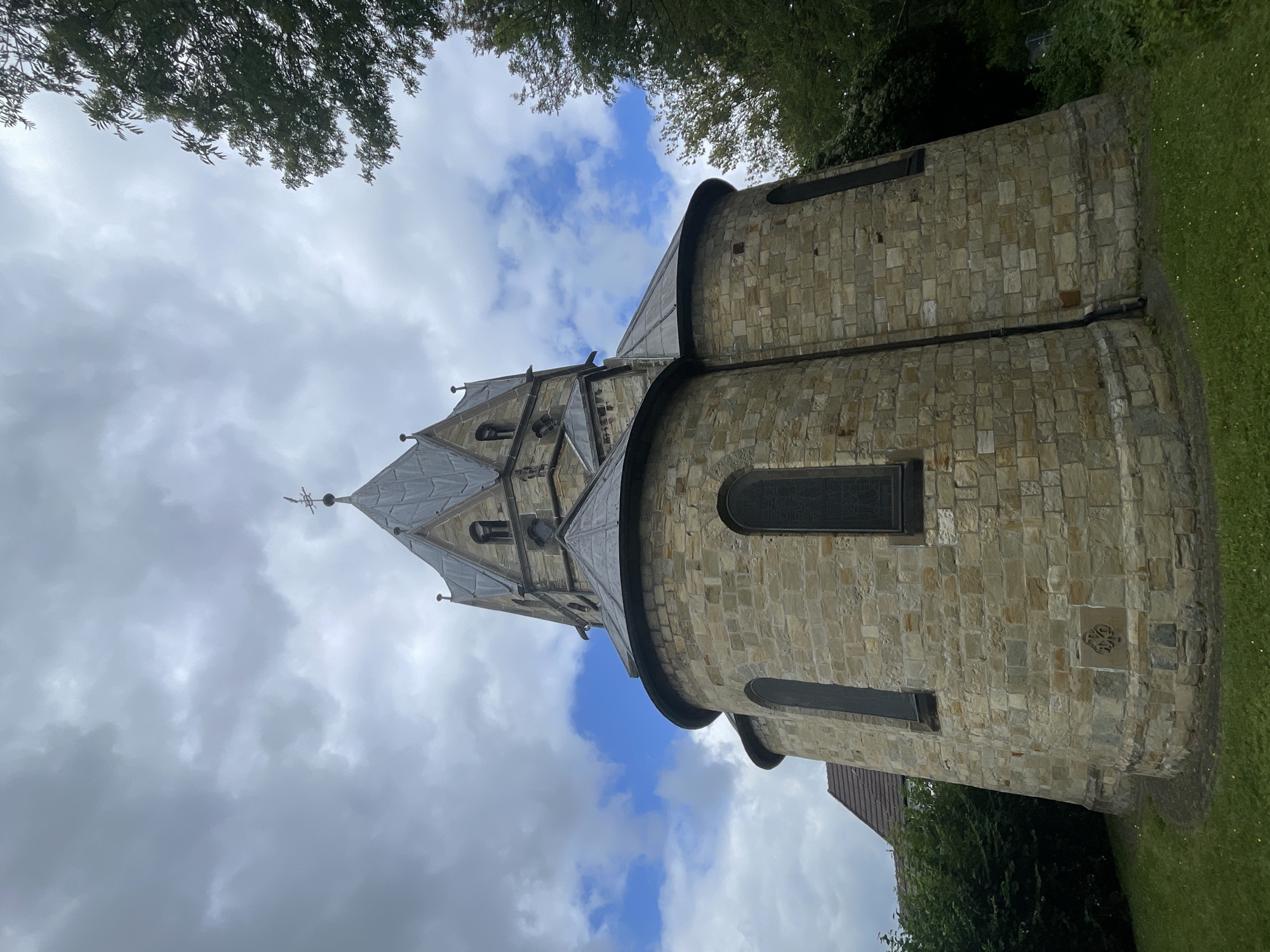The image shows a low-angle, full-shot of a stone church in Salzkotten, Germany. The church has a round base with arched windows, a conical roof, and a tall steeple topped with a weather vane. The sky is cloudy with some patches of blue. Trees and green bushes surround the church. The stone masonry is visible and appears to be of a light brown or tan color. The overall atmosphere is somber due to the overcast sky.
