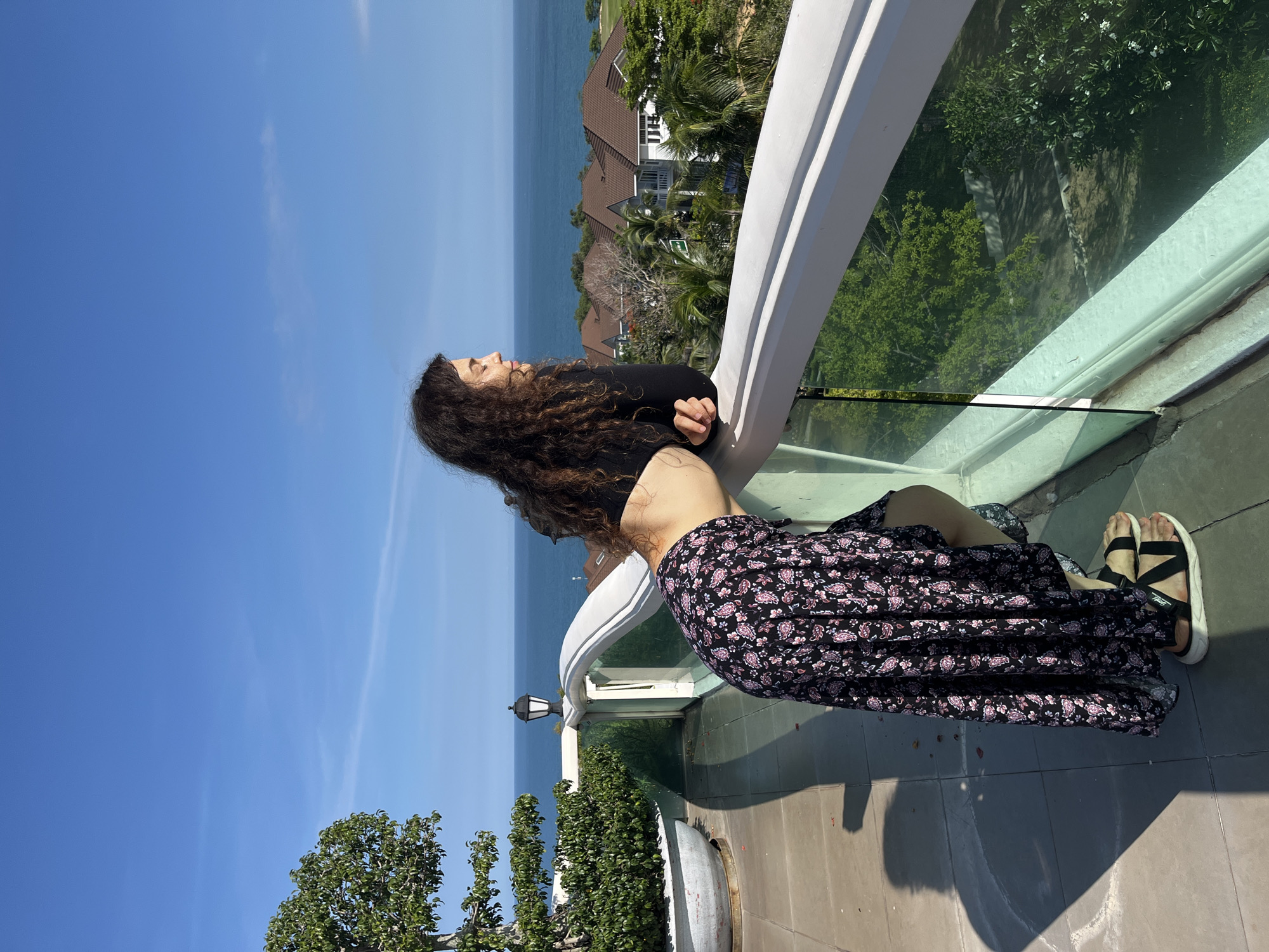 A woman with dark, curly hair stands on a balcony overlooking the sea and a cluster of houses. She is wearing a black crop top and a long, black skirt with a floral pattern in shades of pink and white. Her left arm is resting on the balcony railing, and she is looking out at the ocean. The balcony has a white railing with glass panels. To the left of the woman, a large potted plant with green foliage is visible. The background shows a bright blue sky, the ocean, and several buildings with brown roofs amidst lush green trees and palm trees, indicative of a tropical setting. The scene appears to be located in Phu Thuy Ward, Vietnam, given the architectural style of the houses and the tropical vegetation.