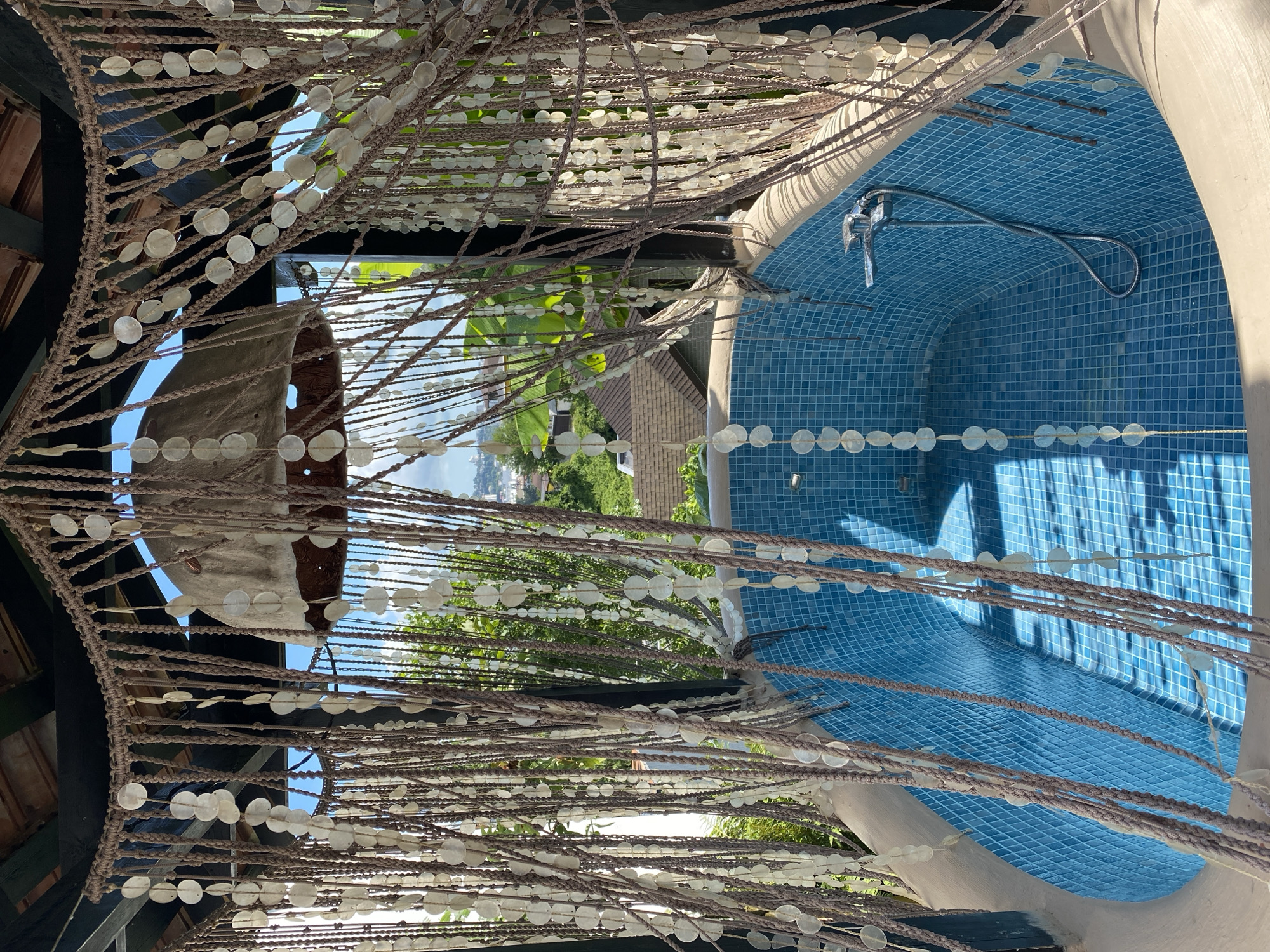 The image captures a close-up view of a beaded curtain made of numerous small, disc-shaped shell or glass beads strung on brown cords. The curtain hangs in front of an outdoor pool area, with the pool itself featuring bright blue mosaic tiles. A shower head is visible attached to the tiled wall of the pool. In the background, through the gaps in the beaded curtain, glimpses of greenery and a building with a tiled roof can be seen, suggesting a warm, sunny environment. The overall impression is of a decorative, perhaps tropical or Mediterranean, outdoor space. The lighting suggests it is daytime, with sunlight casting shadows and highlighting the textures of the beads and tiles. No people or text are visible in the image. The location context provided is Sochi, Russia, which aligns with the possibility of an outdoor pool setting in a region with a temperate climate.