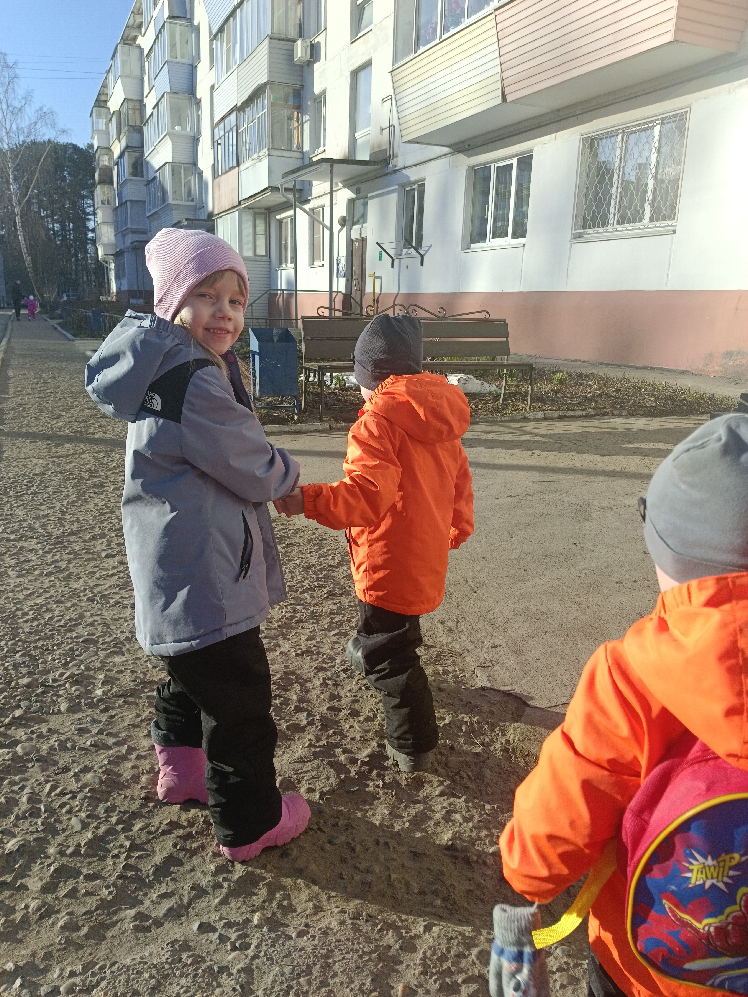 Three children stand in a paved area. A girl in a pink beanie, grey jacket and pink boots looks towards the camera. She holds hands with a child in an orange jacket and black beanie. A third child in an orange jacket, grey beanie and red/blue backpack is partially visible on the right. In the background, there is a multi-story building, a bench, trees, and a clear blue sky. The image was taken in the city of Chaikovsky, Russia.