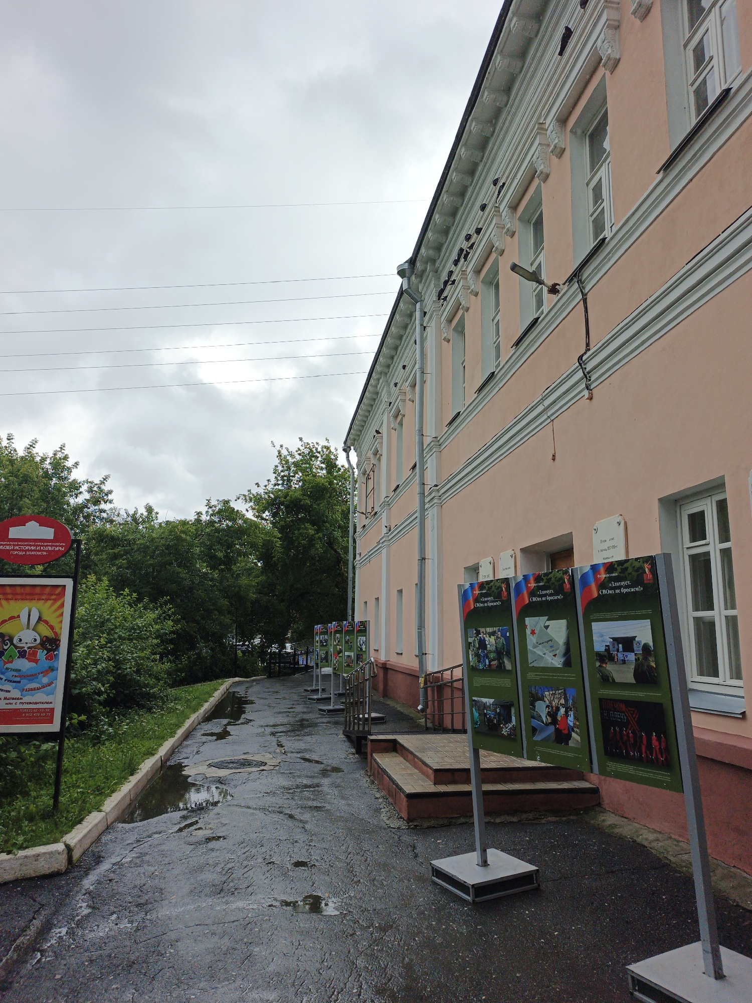 A paved walkway, wet and reflecting the overcast sky, leads alongside a pink building with white trim and decorative architectural details. Trees and lush green foliage line the left side of the walkway. Several display boards stand on the right, featuring patriotic imagery and text in Russian. One board prominently displays the Russian flag and the text "Златоуст. СВОих не бросим" which translates to "Zlatoust. We will not abandon our own." The building appears to be a museum or historical site, indicated by a circular red sign with a silhouette of a building and text that reads "МУЗЕЙ ИСТОРИИ И КУЛЬТУРЫ ГОРОДА ЗЛАТОУСТА" (Museum of History and Culture of the City of Zlatoust). A promotional poster with cartoon characters, including a rabbit, is visible to the left of the museum entrance. The scene suggests an outdoor exhibition or event taking place in front of a cultural institution in Zlatoust, Russia, likely related to current events or historical remembrance. The weather is cloudy and damp, contributing to the wet appearance of the walkway.