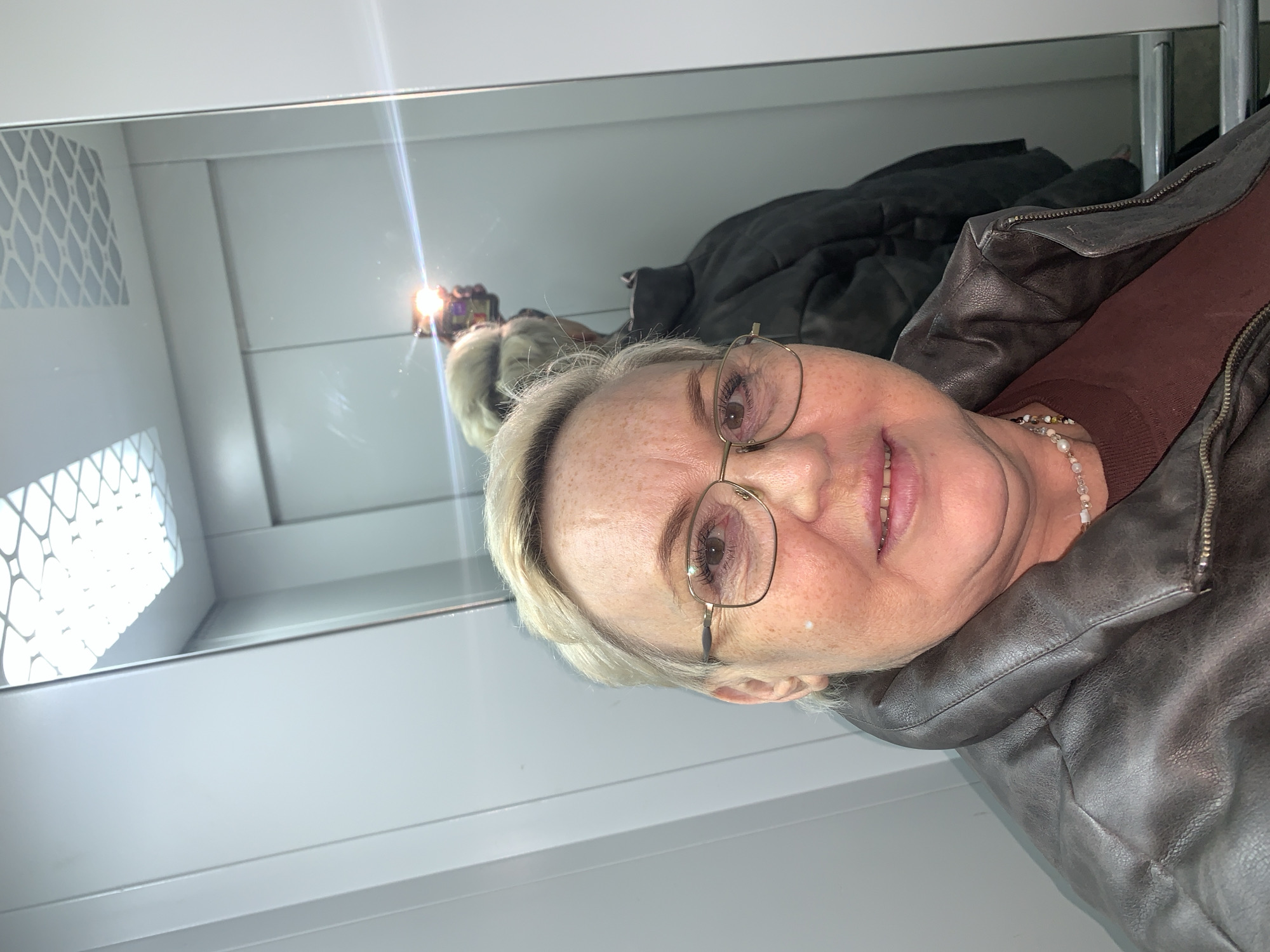 A woman with short blonde hair and eyeglasses is taking a selfie in a mirror. The reflection shows a grey quilted jacket and another person holding a phone. The background appears to be a white room with a patterned section in the upper left. The location is unspecified, no location in Chaikovsky, Russia is mentioned.