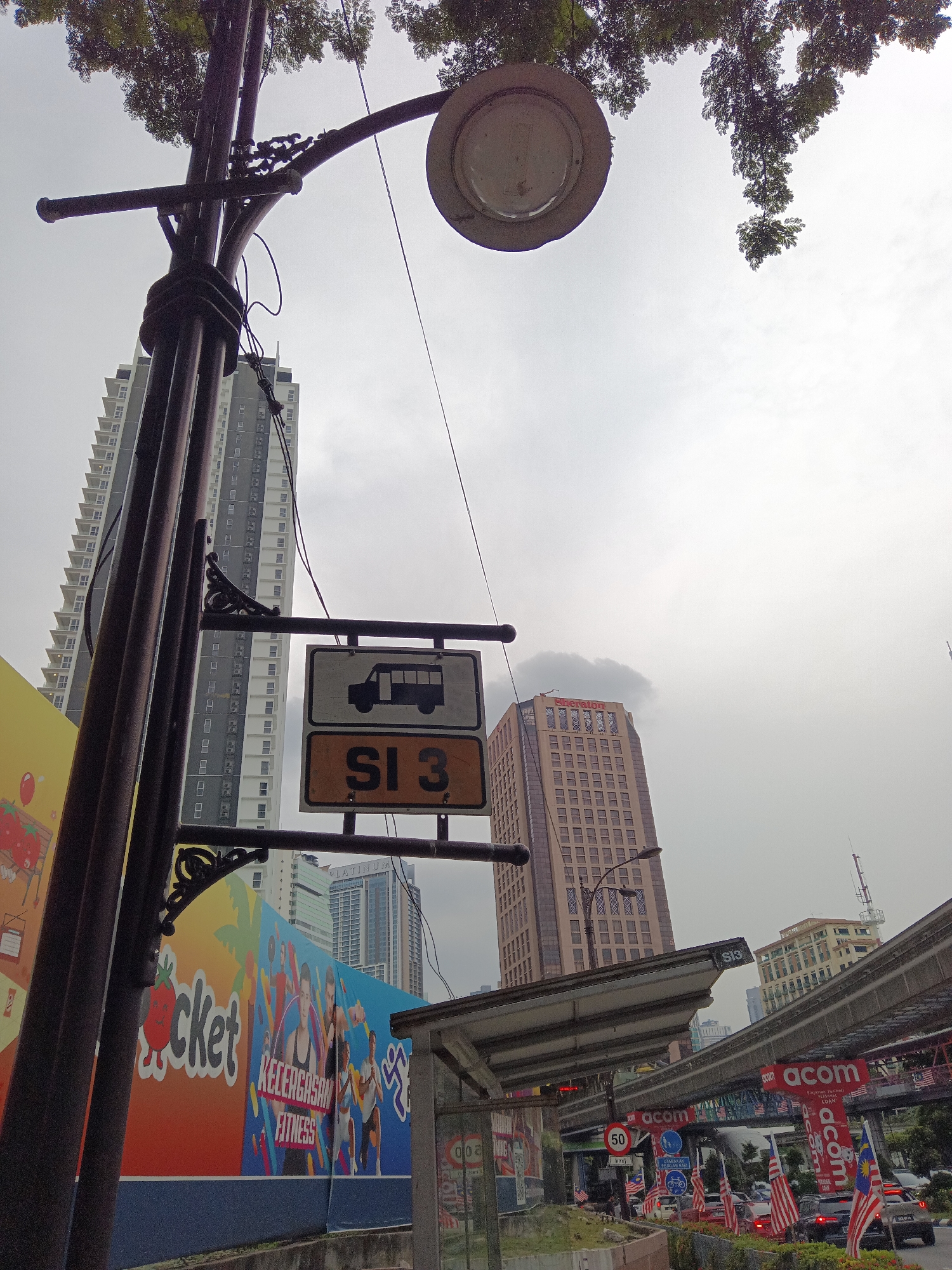 This is a low-angle, full-shot photograph taken outdoors in Kuala Lumpur, Malaysia. The scene is dominated by a tall, dark lamppost in the foreground on the left, adorned with decorative metalwork. Attached to the lamppost is a sign with two panels; the top panel features a black silhouette of a bus on a white background, and the bottom panel displays "SI 3" in black text on an orange background. Behind the lamppost, a tall, modern residential building with many windows stretches towards the sky, its facade a mix of grey and white. To the right of the lamppost, a bus stop shelter with a metal roof is visible. Further in the background, another tall building with a light brown facade and the red "Sheraton" logo is prominent. Overcast skies suggest a cloudy day. In the lower part of the image, there's a vibrant mural advertising a fitness center, depicting people exercising and text that includes "KECERGASAN FITNESS". A raised, elevated train track structure runs across the middle ground of the image. Numerous Malaysian flags are displayed along the roadside, and cars are visible on the street below the elevated track.
