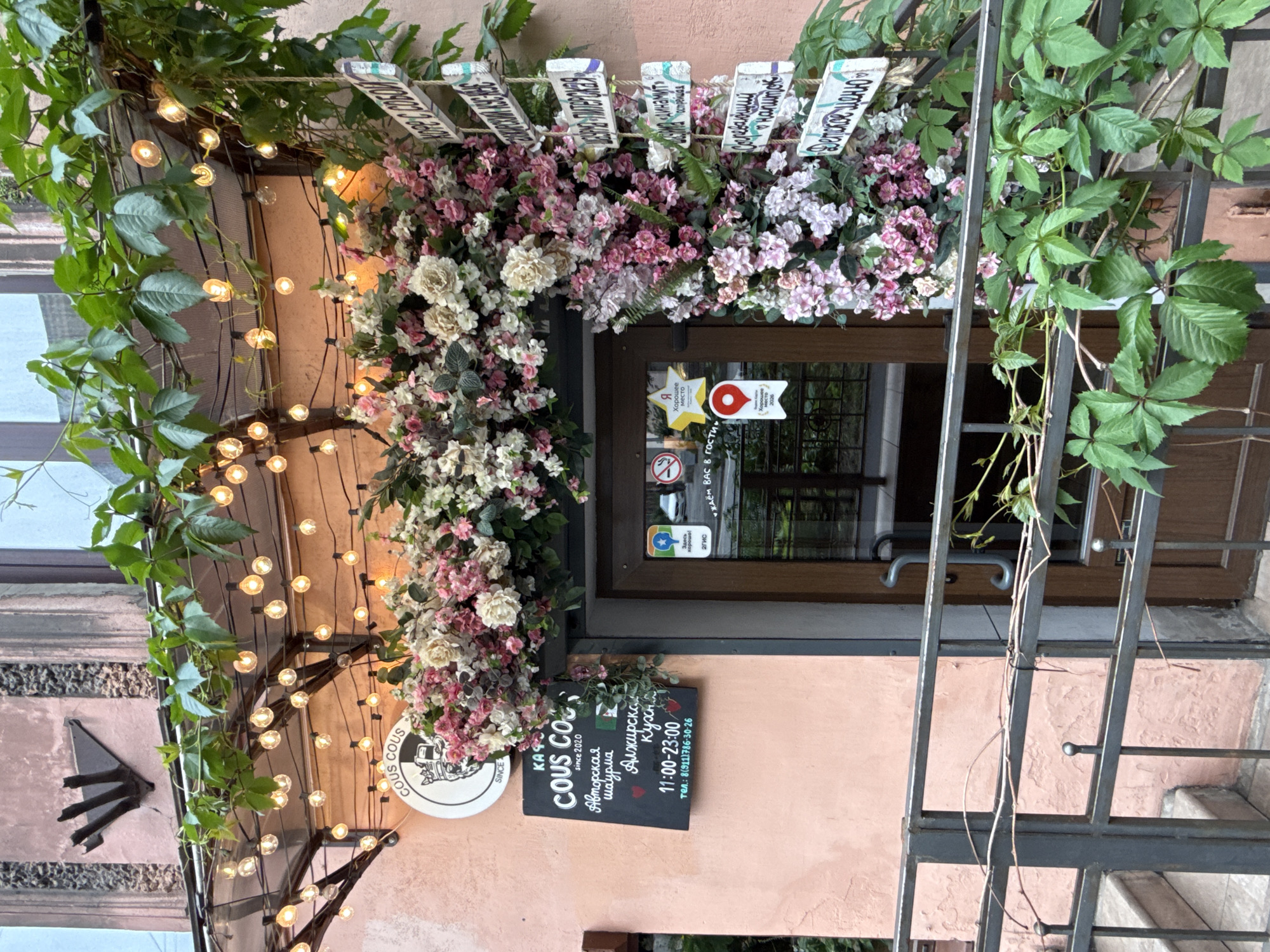 The image depicts the exterior of a cafe in Saint Petersburg, Russia, adorned with abundant floral decorations and string lights. The cafe's facade is painted a pale pink, and a dark brown door is visible. A black chalkboard sign hangs on the wall to the left of the door, displaying the cafe's name, "KOUS KOUS," along with its operating hours (11:00-23:00) and a phone number. Above the door, a canopy is decorated with strings of warm LED lights and lush green vines. To the right of the door, a series of wooden signs are attached to a rope, indicating various menu items or destinations, such as "КОРФ С СОБОЙ" (Coffee To Go) and "ДЕТСКОЕ МЕНЮ" (Children's Menu). A metal railing with decorative finials runs in front of the entrance. The overall impression is a cozy and inviting atmosphere.