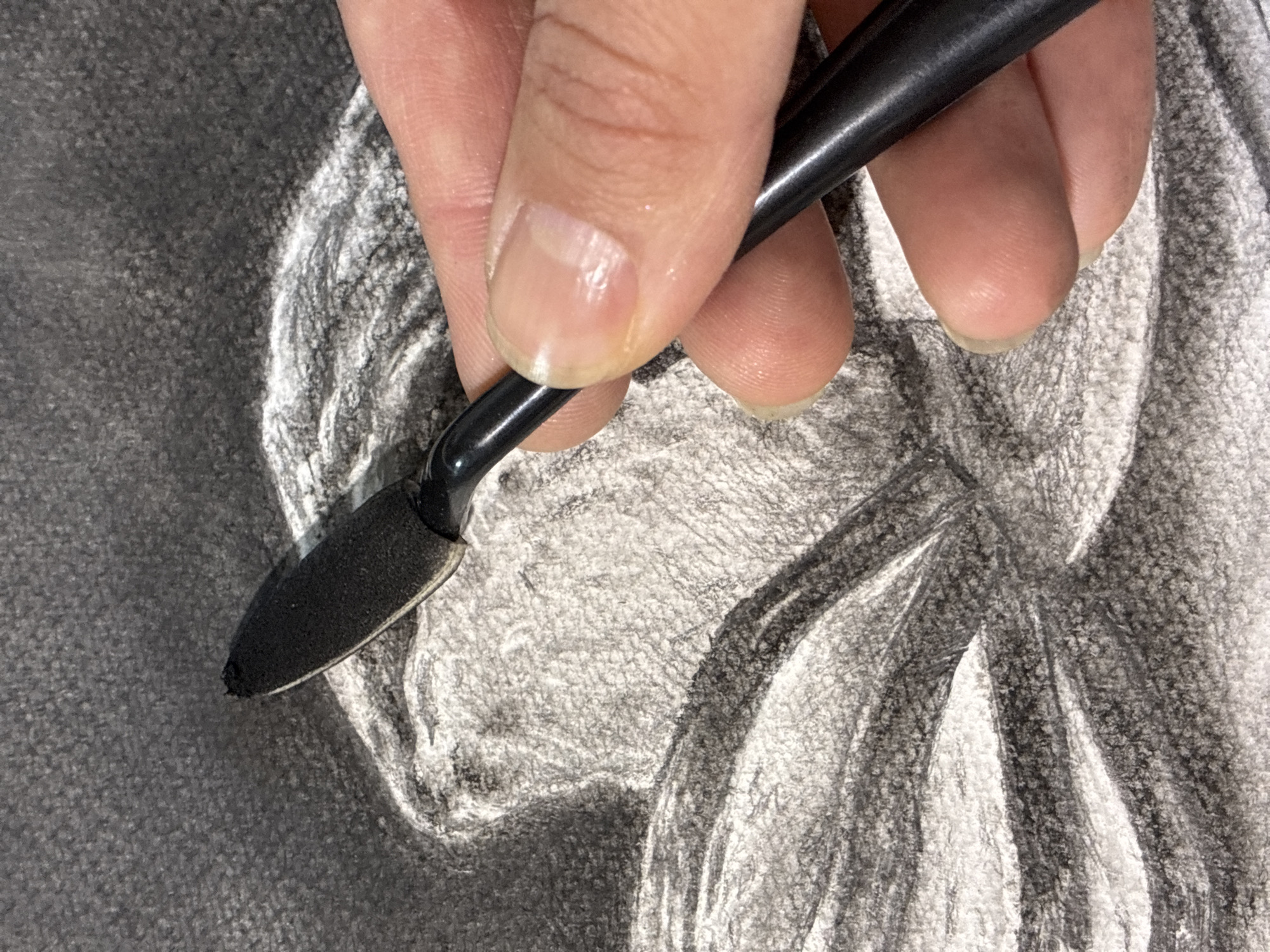 A close-up shot shows a hand holding a black blending tool with a black foam tip against a charcoal drawing. The drawing depicts what appears to be a face, with areas of white and black shading. The hand is positioned to the left of the frame, with the fingers and thumb gently gripping the tool. The blending tool is being used to smudge or soften the charcoal on the drawing. The background is out of focus, but the texture of the paper is visible. The image does not provide any information that would allow for the determination of a location in Bryansk, Russia.