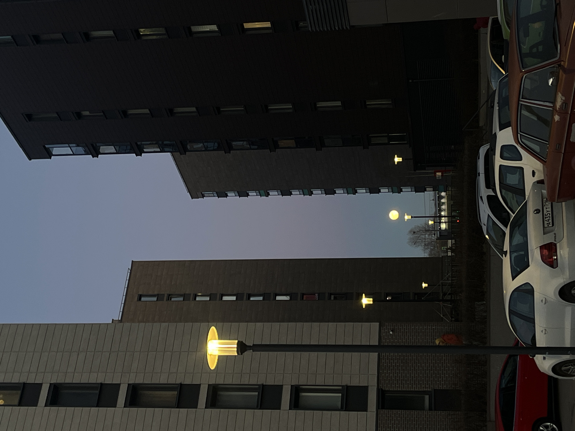 The image shows a street scene in Saint Petersburg, Russia, at twilight. Several multi-story buildings are visible, including one with a dark, monolithic facade. Rows of windows line the buildings. Streetlights are illuminated, casting a warm glow. A row of parked cars occupies the right side of the frame. One car has a visible license plate with Cyrillic letters. A person wearing an orange jacket is visible between two buildings. The sky is a gradient of pale blue to purple.