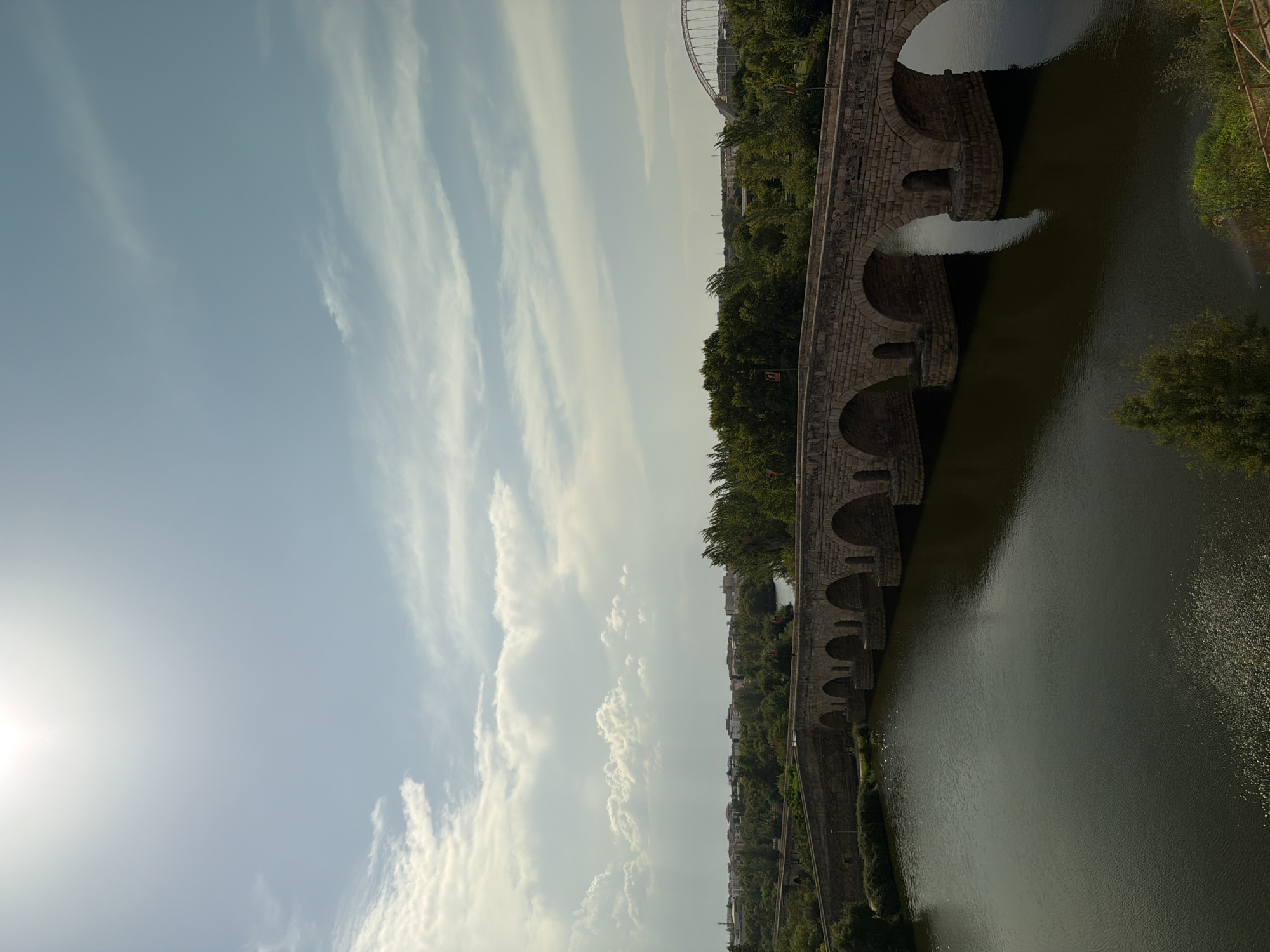 The image shows the Puente Romano bridge in Mérida, Spain. The ancient Roman bridge spans the Guadiana River. The stone bridge has numerous arches and is lined with greenery on both banks of the river. The sky is partly cloudy with the sun shining brightly. In the background, there is a modern bridge with a distinctive arch. The scene is captured from an elevated viewpoint looking across the bridge and river.