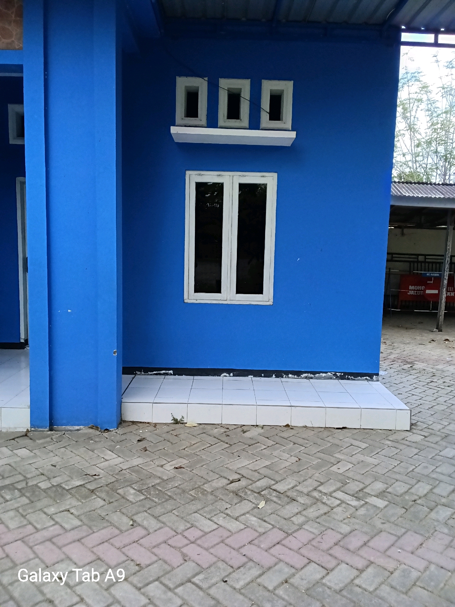 This image depicts a bright blue building with white-trimmed windows and a white ledge. The building's facade is smooth and painted. There are three small, rectangular windows stacked vertically on a white ledge, and one larger, rectangular window below them. The building is situated on a paved area made of interlocking bricks. In the background, a partially visible structure with a corrugated metal roof and metal fencing can be seen. Signs with red lettering are visible on the fence, including text that appears to read "MOHON JALUR" and "ILL ANT". The setting is outdoors, with some foliage visible behind the building, suggesting a location in Karangan, Indonesia. The lighting indicates it is daytime. The image has the text "Galaxy Tab A9" in the lower left corner.
