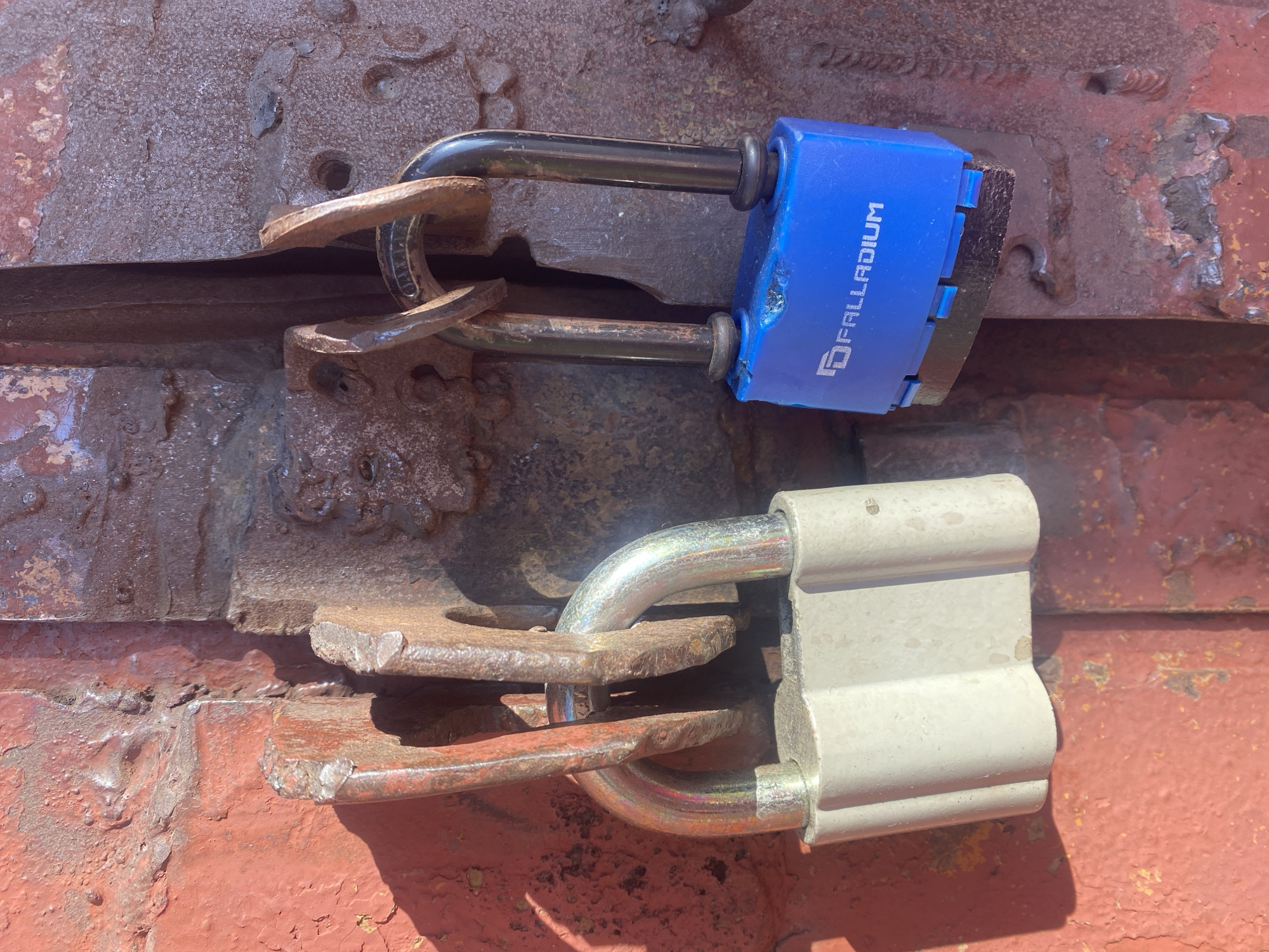The image displays two padlocks securing a closure on a heavily rusted, red-brown metal surface, likely a door or gate. The scene is illuminated by bright daylight, casting distinct shadows from the locks onto the textured background.
The upper padlock is blue with a black shackle. The brand name "PALLADIUM" is clearly visible in white capital letters on its side. This padlock is fastened through two rusty, weathered metal staples or hasps.
Below it, a second padlock, lighter in color (possibly gray or off-white) with a metallic shackle, is similarly secured through another set of rusty metal staples.
The metal surface itself shows extensive signs of age and wear, with peeling paint, deep rust, and an uneven, pitted texture. This suggests a rugged, outdoor, and possibly industrial or storage context. No human subjects are present, and no activity is taking place; the locks are static and functional. The overall impression is one of utility and resilience against the elements. The location is implied to be Novokuznetsk, Russia.