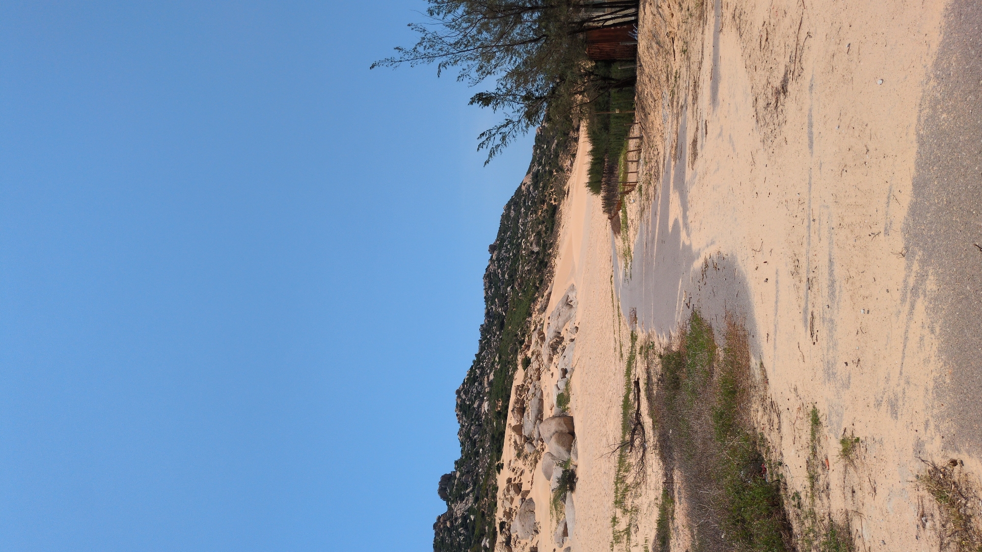 The image shows a landscape in Phuoc Dinh, Vietnam. The scene includes a sandy hill covered with rocks and some vegetation. A dirt road runs alongside the hill, transitioning into a paved surface on the right side of the image. Trees are visible at the top of the hill, near what appears to be a wooden fence. The sky is clear and blue.