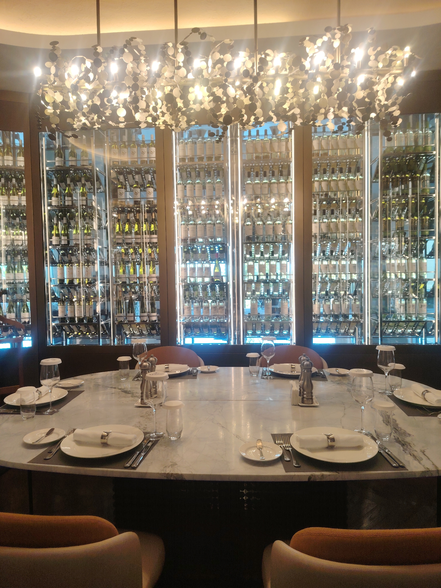 A high-end dining establishment is depicted in this image, likely a restaurant. The scene is centered around a large, oval marble table set for a formal meal. Each place setting includes a white plate with a folded white napkin secured by a decorative ring, cutlery, a water glass, and a wine glass. Salt and pepper grinders are present in the center of the table, along with a tissue dispenser. In the background, a prominent wine cellar stretches across the wall, filled with numerous bottles of wine organized on illuminated shelves. A unique, artistic chandelier hangs above the table, casting a warm glow on the scene. The chairs around the table are upholstered in a neutral fabric, some with hints of orange or tan. The overall ambiance suggests a sophisticated and luxurious dining experience. The image does not contain any information or visual cues to suggest a location in Konigil, Uzbekistan.
