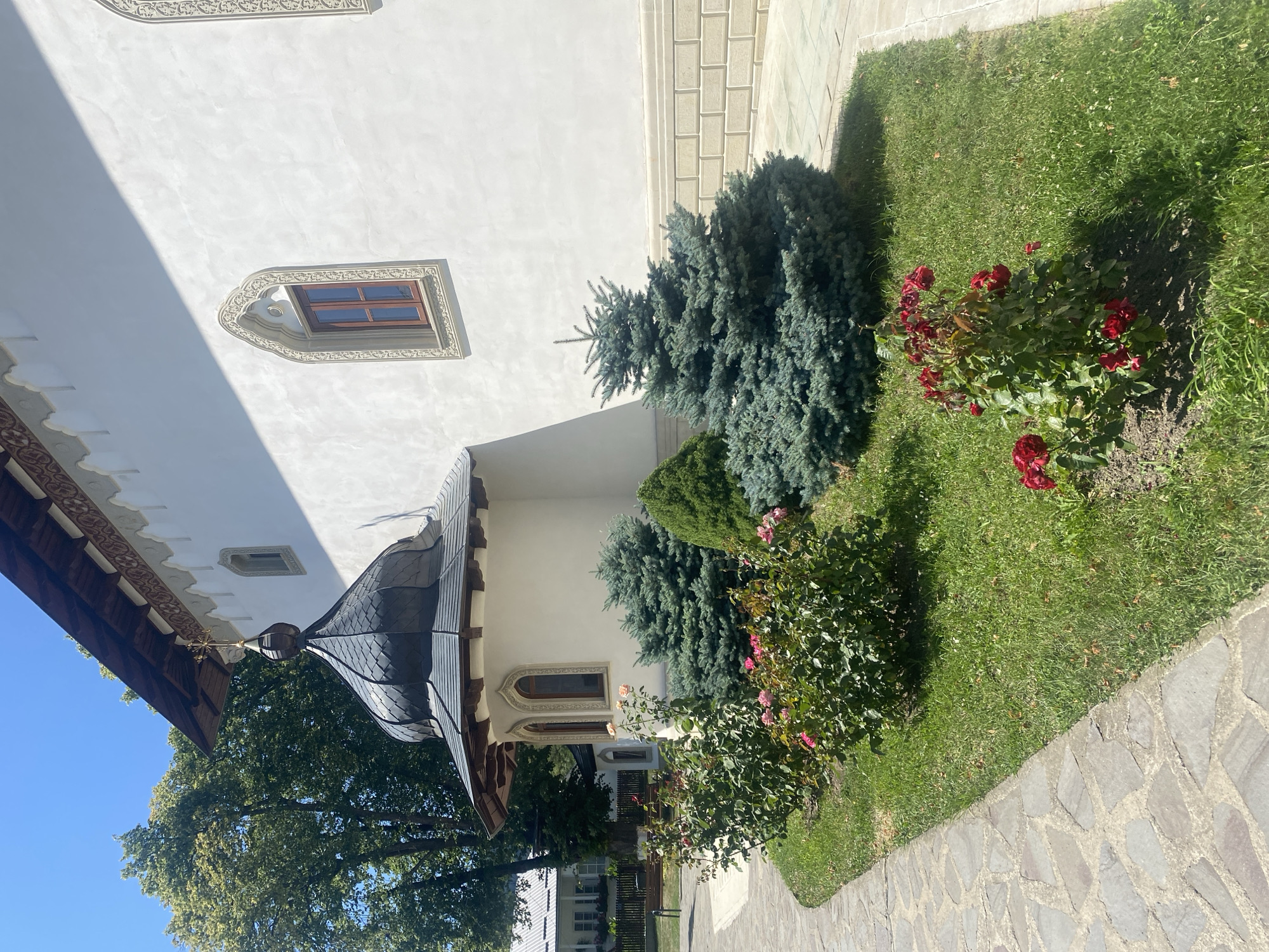 The photograph shows a section of a white-walled building with a dark, domed roof and a golden cross at its apex. The building features ornate windows with dark wooden frames. In front of the building, a path made of irregularly shaped stones curves to the left. Adjacent to the path is a lawn with several evergreen shrubs and rose bushes in bloom. One rose bush displays red flowers, and another has lighter-colored roses. The scene is bathed in bright sunlight, casting shadows. In the background, a large tree with green foliage stands to the left, and to the far left, the roofline of another building is visible, suggesting a larger complex. The location is identified as Filioara, Romania.