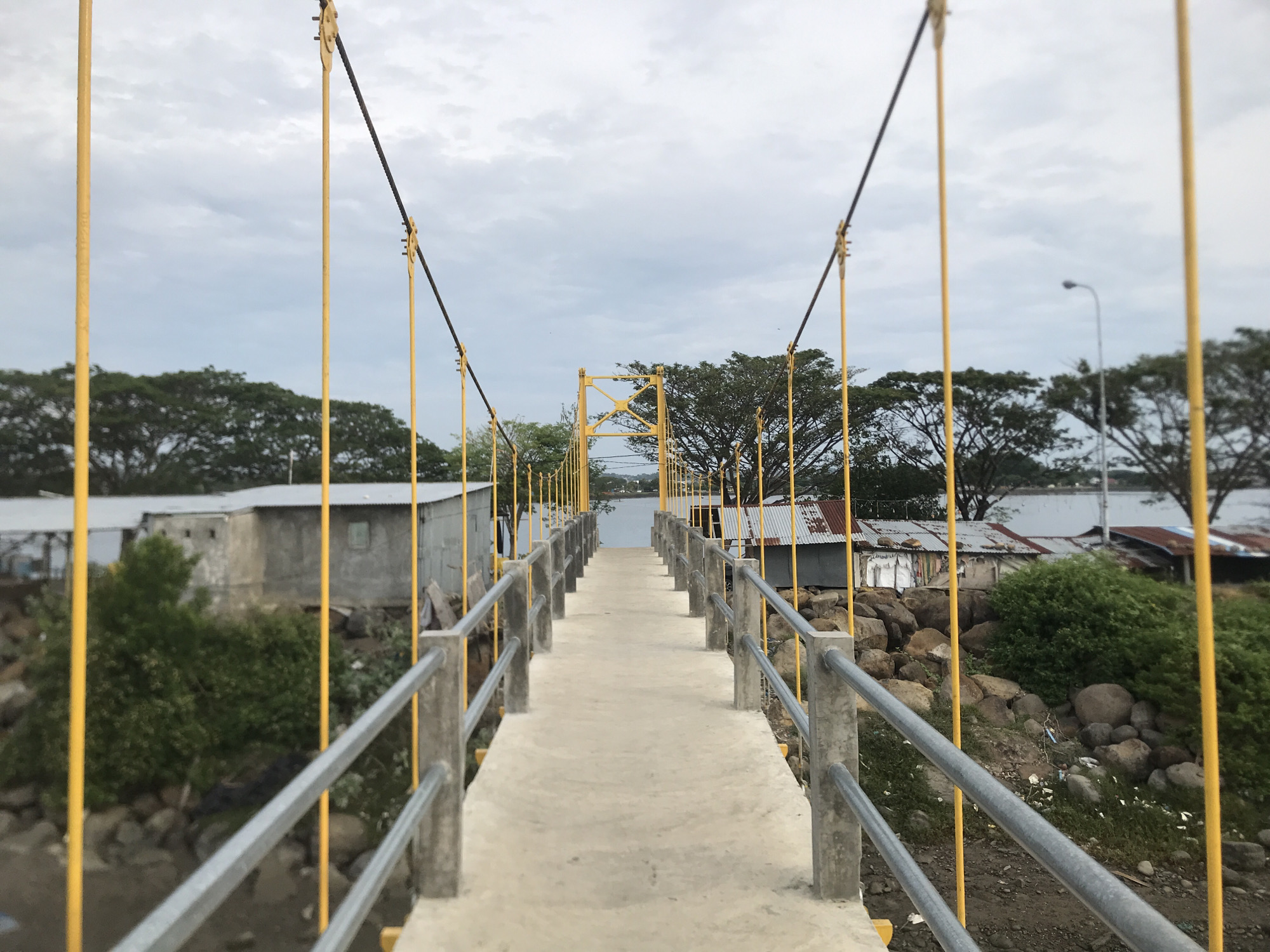 This is a medium shot, eye-level, outdoor photograph taken on an overcast day, looking down a pedestrian suspension bridge. The bridge is made of concrete and has grey metal railings supported by concrete posts. Bright yellow poles line the sides of the bridge, supporting cables that ascend to a central yellow metal support structure in the distance, indicating the bridge's suspension design.
On either side of the bridge, there are a few modest, weathered structures with corrugated metal roofs, some appearing to be dwellings or small businesses. Vegetation, including bushes and trees, grows around these structures and along the bank. The ground on the right side of the bridge is rocky and uneven, with some scattered debris. A street lamp is visible on the right, further down the embankment.
The bridge leads towards a body of water, likely a river or bay, under a sky filled with grey clouds. The overall scene suggests a functional pedestrian crossing in a relatively modest or developing area. This type of infrastructure is common in coastal or riverine communities, and the visual elements are consistent with areas in Lhokseumawe, Indonesia, which is known for its coastal geography and port facilities. Specifically, this appears to be a pedestrian bridge over a small waterway or channel within the city. There are no people visible in the photograph.