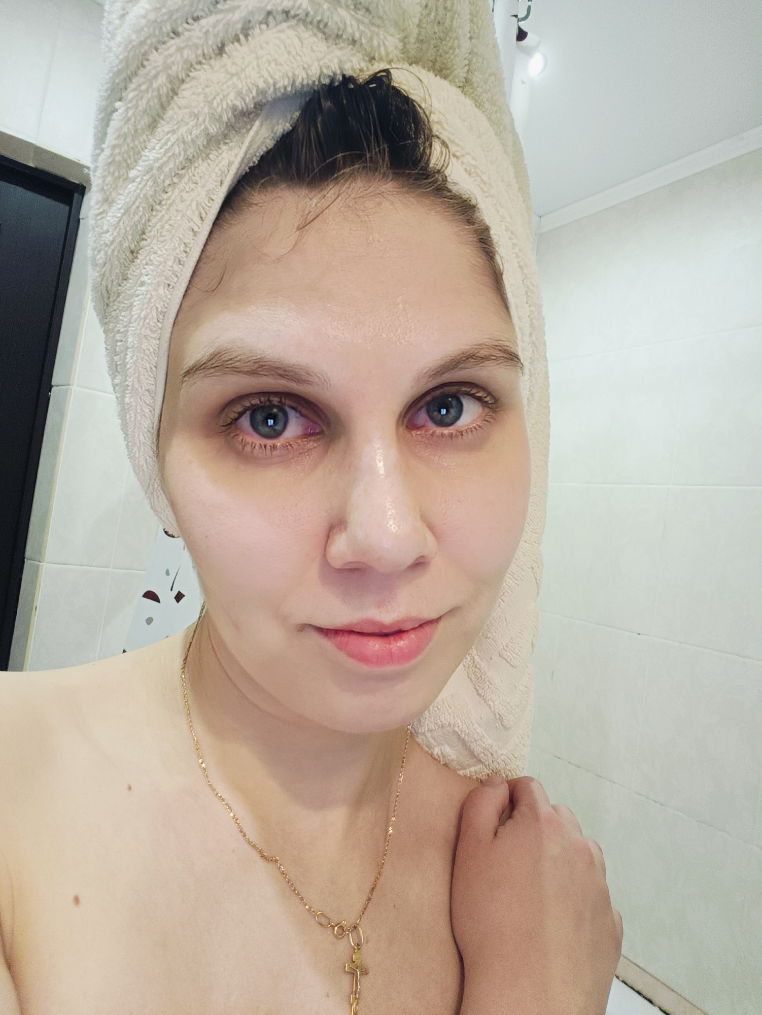 A woman with a fair complexion and blue-grey eyes is featured in a close-up, chest-up portrait. She looks directly at the camera with a soft, closed-mouth smile. A light-colored, textured towel is wrapped around her head, indicating her hair is wet, possibly after a shower or washing her face. Her skin appears moisturized or slightly dewy, with a noticeable sheen on her forehead and the bridge of her nose. There is a slight redness visible on her lower eyelids.
She is wearing a thin gold chain necklace with a small cross pendant. Her bare shoulders are visible in the lower part of the frame. The background suggests an indoor setting, likely a bathroom, with white tiled walls. A dark door is partially visible on the left side, and a light fixture can be seen on the ceiling in some of the cropped views. The lighting is artificial. There is no visible text in the image.