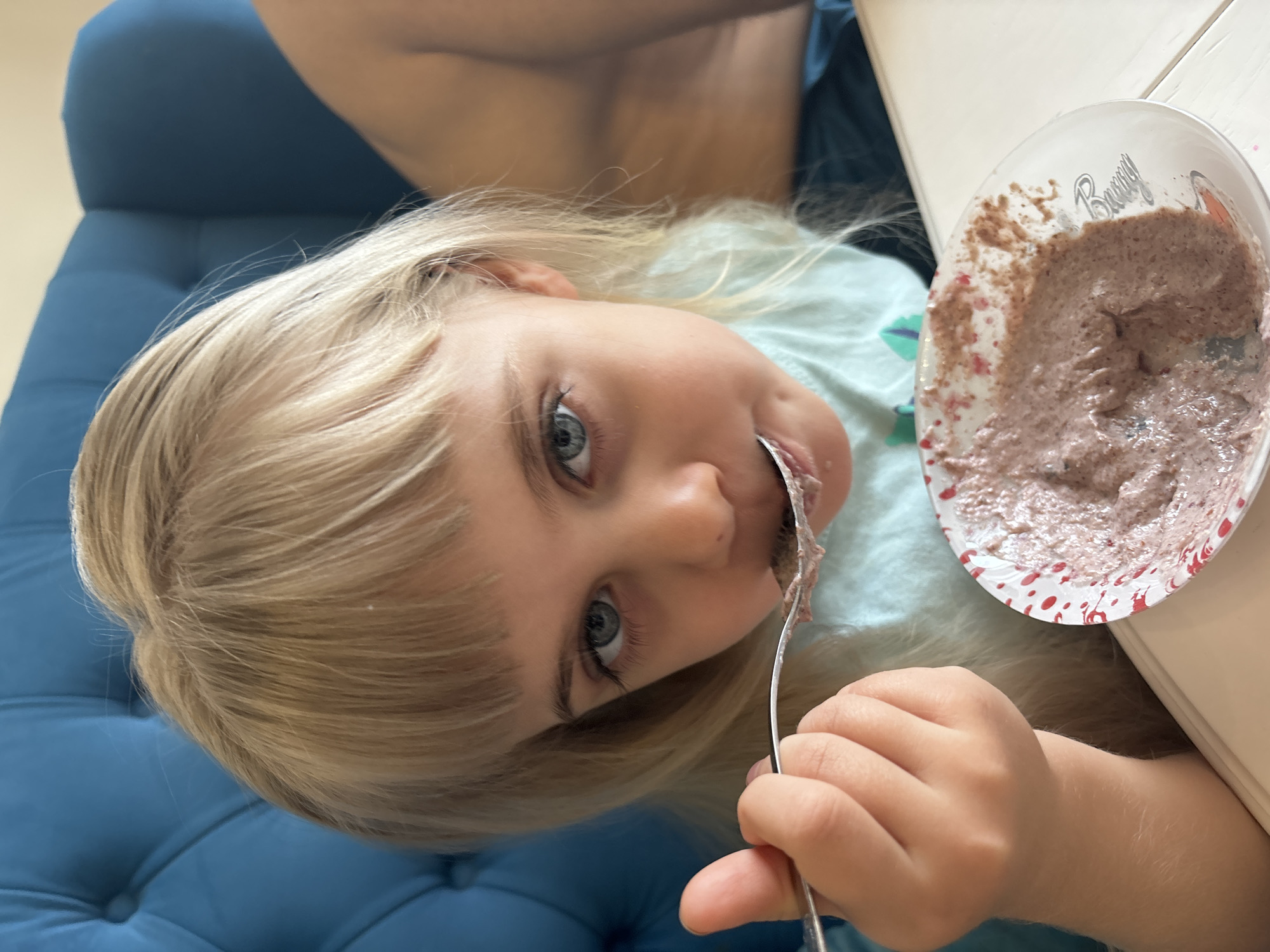 A young child with blonde hair and blue eyes is eating something from a bowl with a spoon. The bowl is white with red splatters and the word "Bunny" on it. The child is sitting on a blue padded chair or couch, next to a white table. The location is unknown.