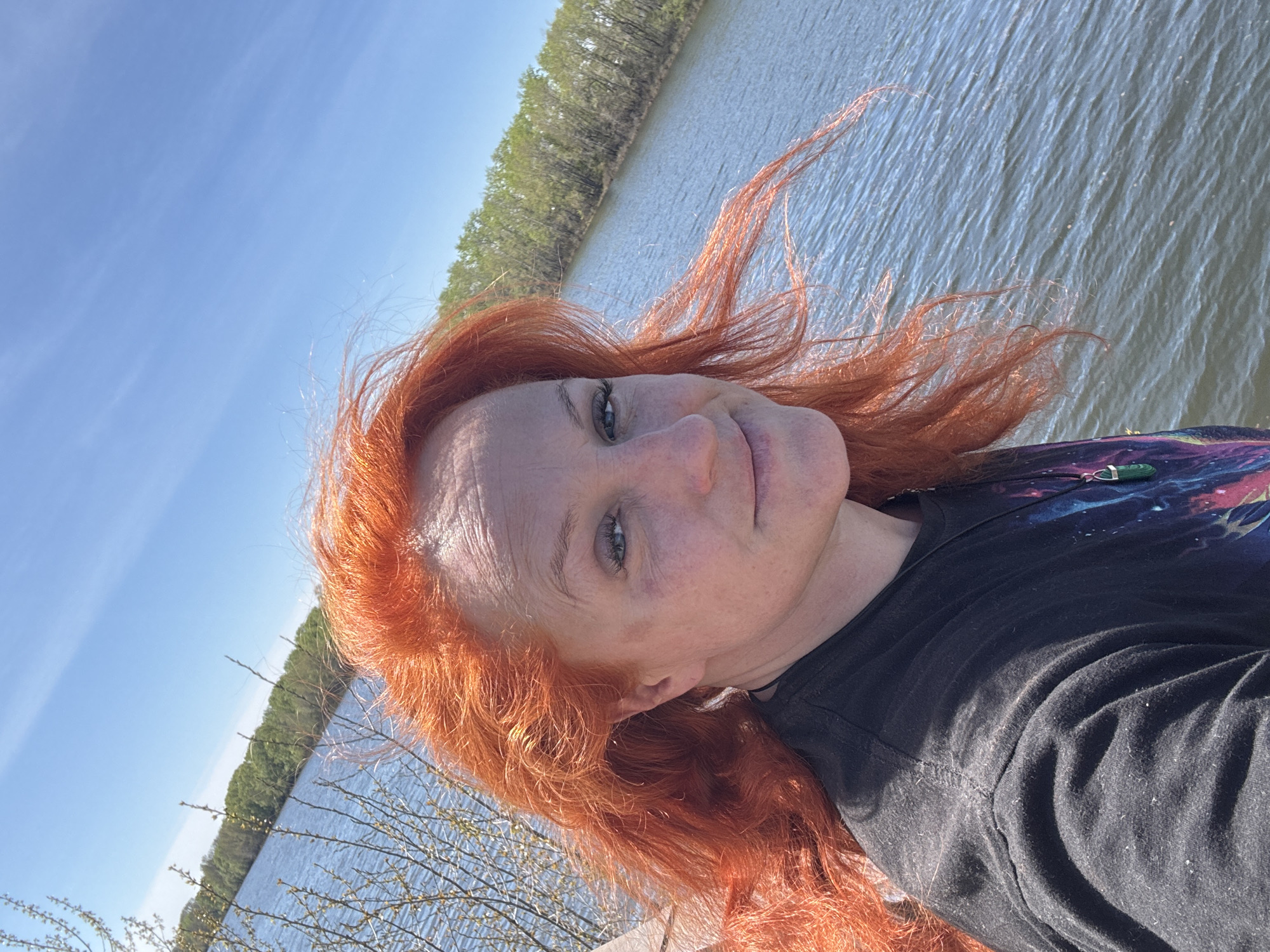 A person with bright red hair is pictured in a selfie taken outdoors on a sunny day. The background includes a body of water, likely a river or lake, with a tree-lined shore in the distance. The sky is a clear blue. The person is wearing a dark-colored shirt and a necklace with a green stone pendant is visible. The photograph was taken on the banks of the Kama River, near the city of Chaikovsky, Russia.