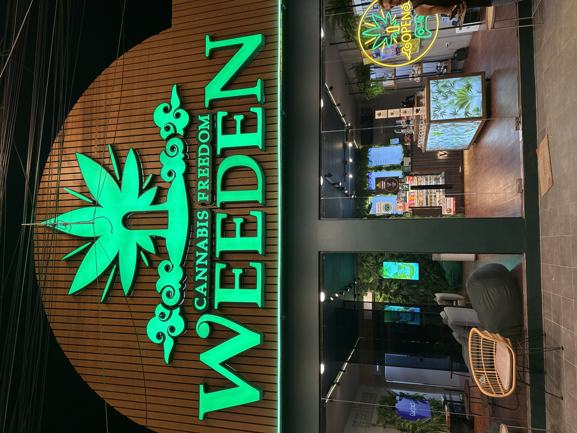 The image shows the exterior of a cannabis shop called "WEEDEN CANNABIS FREEDOM" located in Baan Bo Phut, Thailand. The shop's facade features a large, vertically oriented green neon sign with the shop's name and a cannabis leaf logo. The sign is mounted on a wooden paneled wall.
Through the glass storefront, the interior of the shop is visible. The shop appears to be modern and well-lit, with displays showcasing cannabis products and plants. There are several screens displaying greenery and product information. Comfortable seating areas with beanbag chairs and a wicker chair are visible, suggesting a space for customers to relax.
In the background, a person is partially visible, holding a phone and seemingly looking into the shop. Another person is seen standing near the entrance on the right side of the image. The time of day appears to be evening or night, as indicated by the dark sky and the illuminated neon sign. The overall impression is that of a contemporary cannabis dispensary.
Visible text includes:
- "WEEDEN CANNABIS FREEDOM" on the main sign.
- "OPENS" with a clock symbol on a neon sign inside the shop.
- "We're not 'pot-heads' but we do love to smoke" on a sign near the entrance.
- "TRIPADVISOR" logo on a sign.
- Some text on screens inside the shop, including what appears to be product names or menus.
- "Choczerd" on a purple sign near the bottom left.