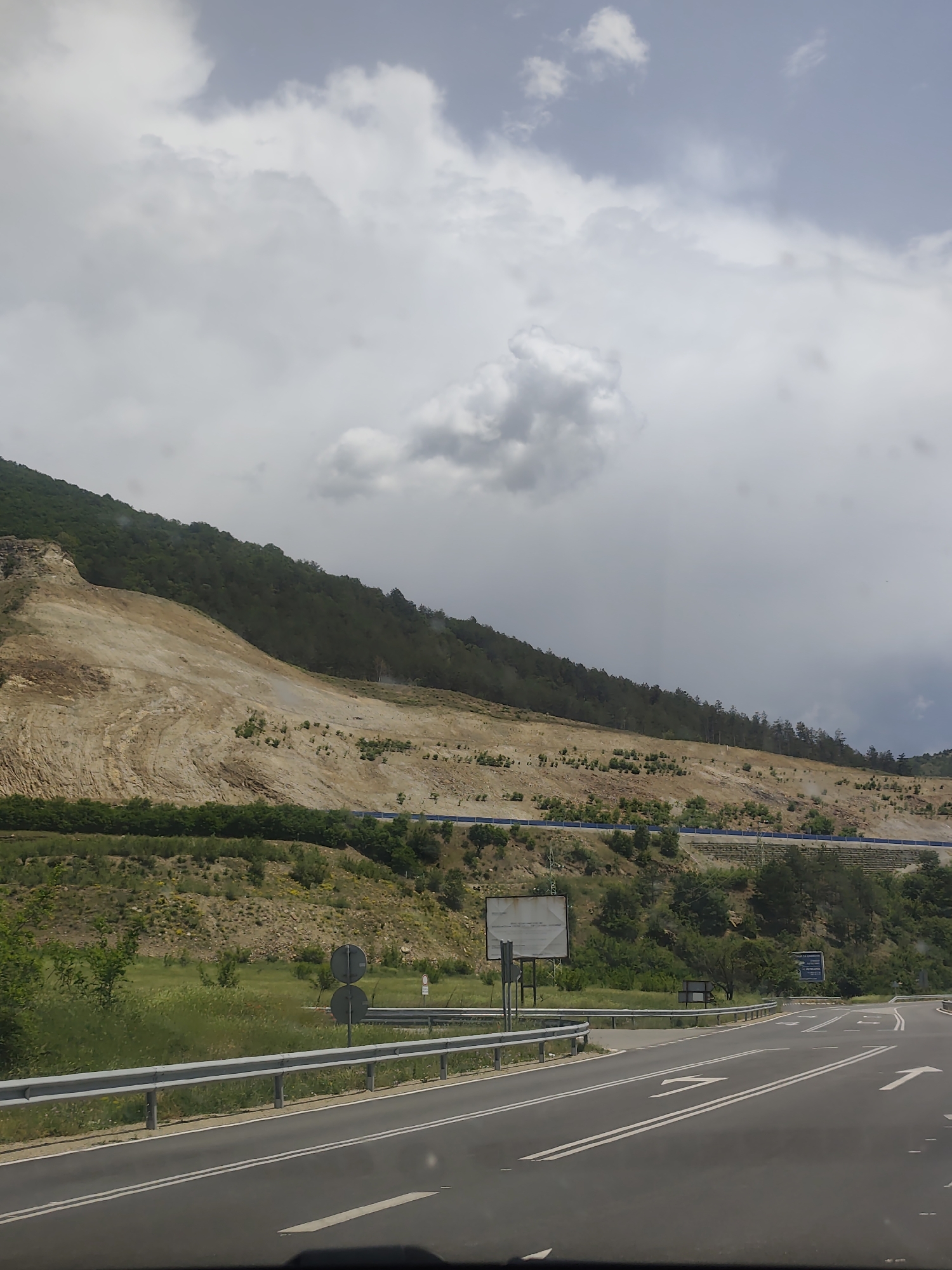 The image shows a road scene near Jeleznitsa, Bulgaria. In the foreground is an asphalt road with white lane markings and directional arrows, bordered by a metal guardrail. Beyond the guardrail is a grassy area with sparse vegetation and road signs. In the midground, a deforested hillside is visible, revealing layers of earth and rock, with a line of trees at the top. A blue safety barrier runs along the slope. The sky is overcast with gray clouds.