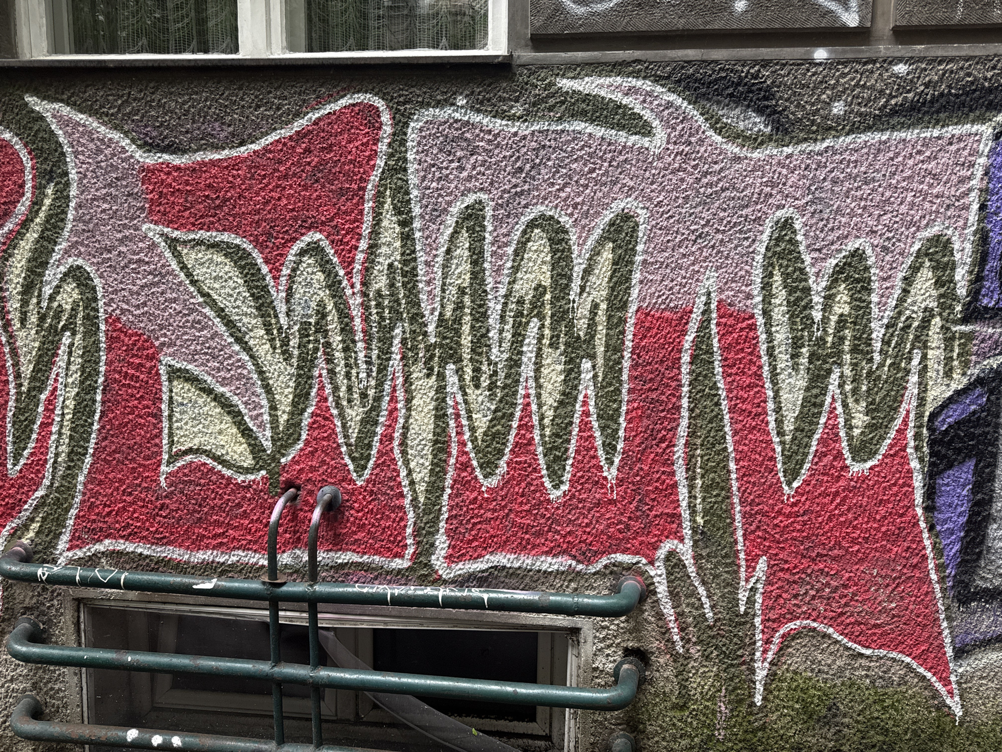 The image depicts graffiti on a textured wall in Belgrade, Serbia. The graffiti features red, purple, and gray sections outlined with white and green lines. Below the graffiti, there is a partially visible window secured by green pipes. A second window can be seen at the top of the image.