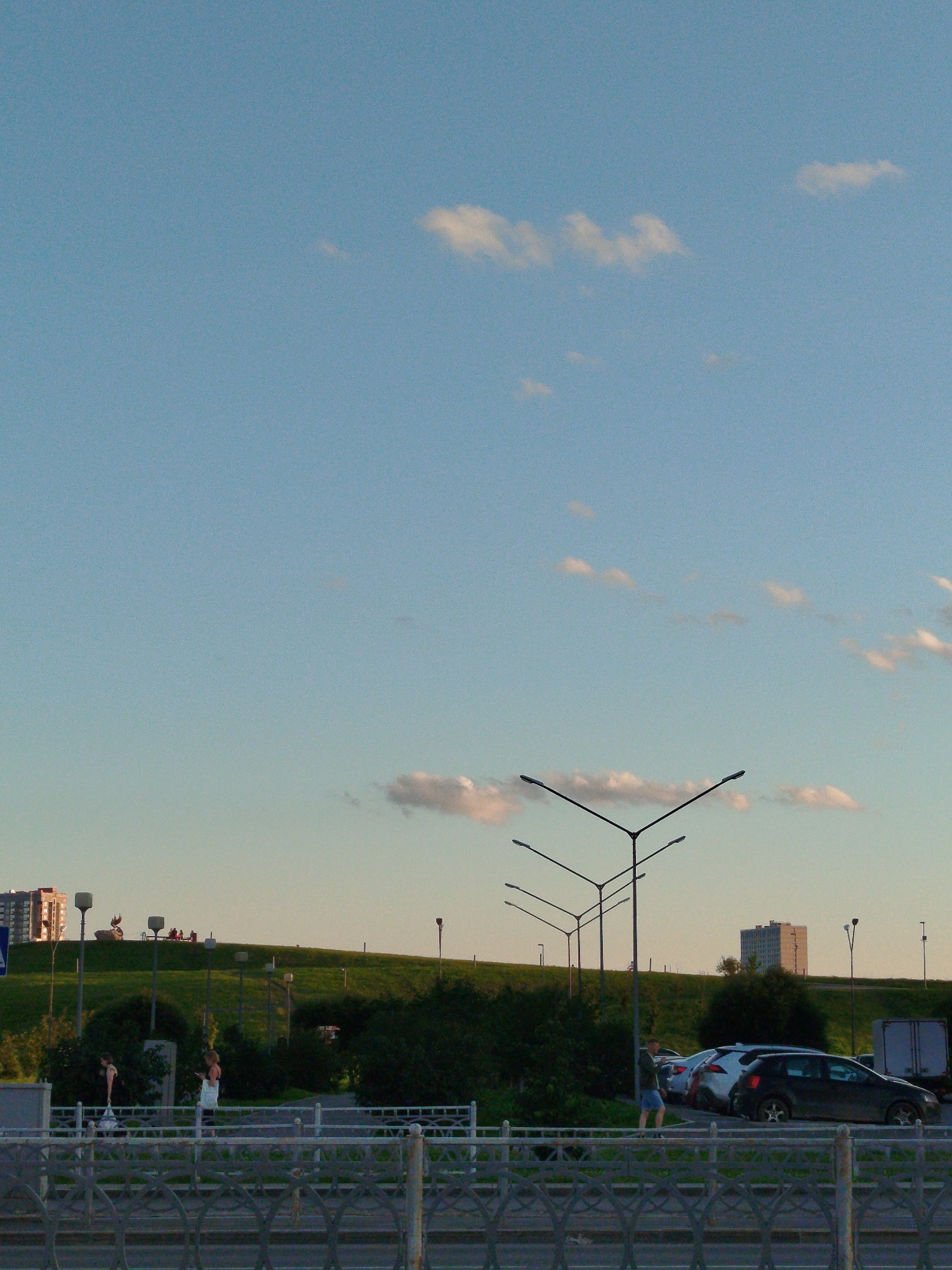 This image depicts a view of the Shartash Stone Hills natural park in Yekaterinburg, Russia, on a clear, bright day with a few scattered clouds. In the foreground, a metal railing runs horizontally across the frame, obscuring the lower portion of the scene. Beyond the railing, a paved area and a walkway are visible. Two women are present on the walkway; one is wearing a white dress and appears to be holding a phone, while the other, in the distance, is wearing a pink dress and standing next to a lamppost.
Further in the background, a green, grassy hill dominates the landscape. Several lampposts are scattered across the hill, and a group of people are seated on top. Two buildings are visible on the horizon: a multi-story building on the left and a taller, light-colored building on the right. A parking lot with several cars is situated at the base of the hill on the right side of the image. The sky above is a pale blue, transitioning to a warmer hue near the horizon.
