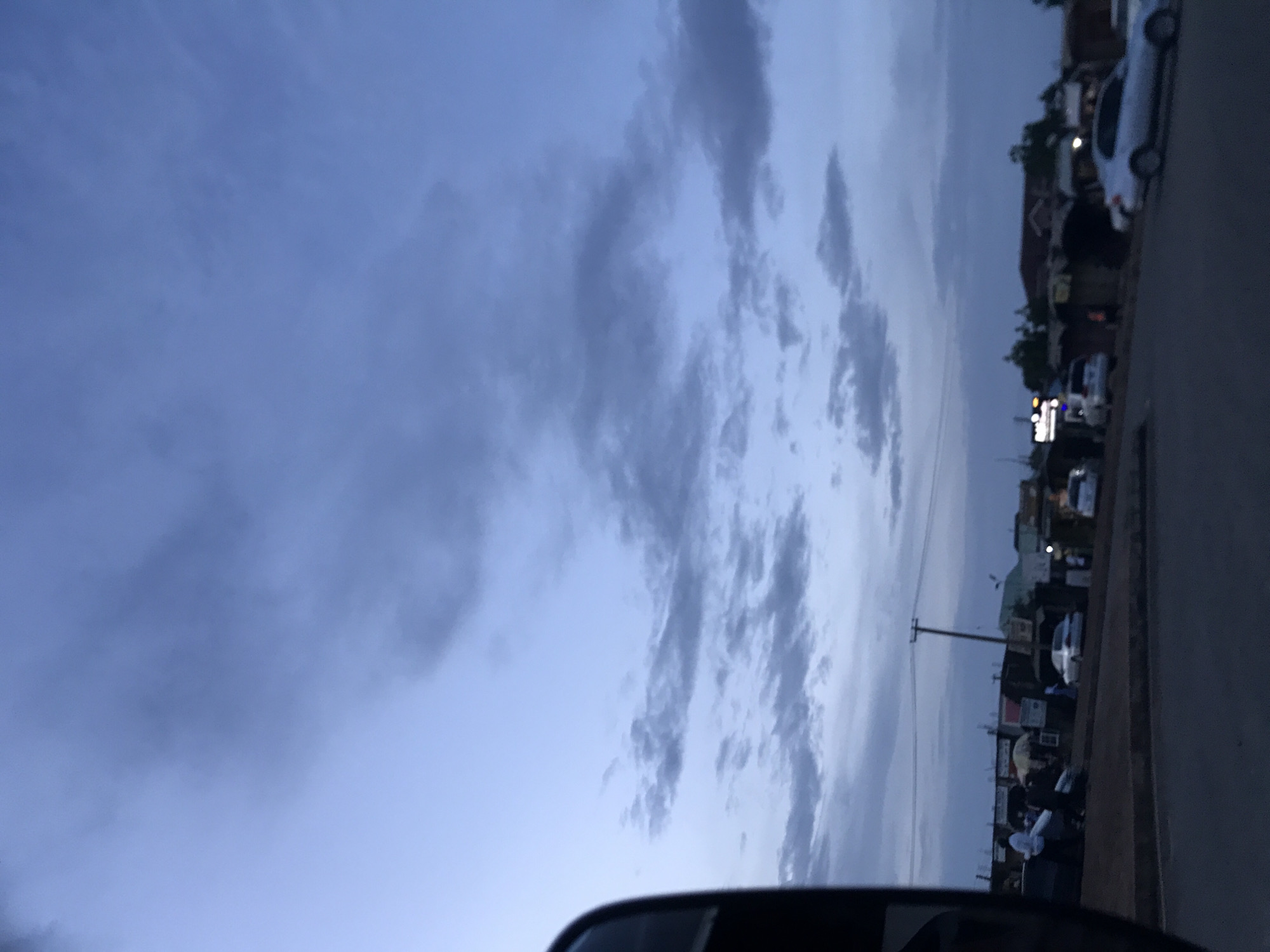 The image depicts an outdoor urban scene in Rafin Par, Nigeria, observed during twilight, either at dusk or dawn. The upper two-thirds of the frame are dominated by a blue-grey sky with scattered, darker stratocumulus clouds. The ambient light is low, casting a subdued tone over the entire scene.
In the foreground, a dark, curved object, consistent with the frame of a vehicle's side mirror or interior, partially obstructs the view on the left. The lower third of the image features a paved road or street that curves slightly towards the right. Along the right side of the road, a collection of low-rise commercial structures or stalls with corrugated metal roofs and various signs are visible. Several people are present in this area; one individual wearing a white top stands near a parked vehicle on the left, and another person appears to be operating or standing near a motorcycle or scooter.
Multiple vehicles are present on and adjacent to the road. At least two light-colored sedan cars are distinguishable, one parked on the right and another moving or parked further along the road. Overhead utility lines and a wooden utility pole are visible in the mid-ground, indicating basic infrastructure. Distant buildings and faint artificial lights are discernible in the far background, further obscured by the low light conditions. The scene suggests a period of reduced activity, characteristic of early morning or late evening in a commercial district.