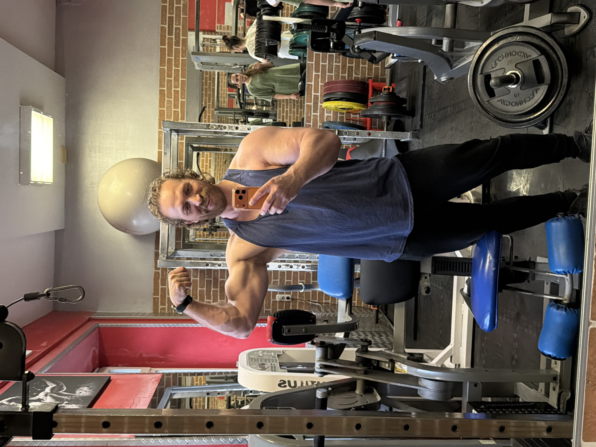 A man is in a gym, flexing his arm and holding a phone in the other. There is gym equipment surrounding him. The background shows a mirror reflecting the man, other gym-goers, and additional equipment. The gym has gray and brick walls. The city and country are not discernible.