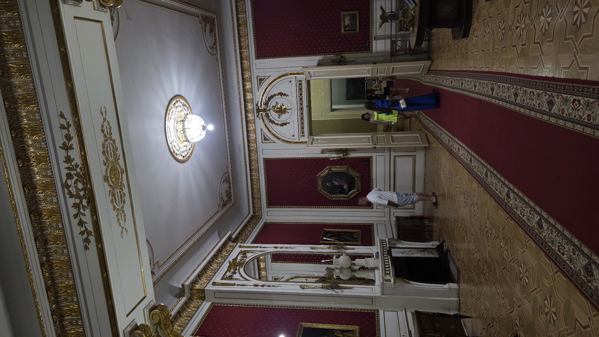 The image depicts the opulent interior of an art museum or palace, possibly the Lviv National Art Gallery in Lviv, Ukraine. The room features high ceilings adorned with intricate gold ornamentation and a large chandelier. The walls are predominantly covered in rich red fabric with a subtle pattern, accented by white panels and more gold detailing. Several framed paintings are hung on the walls, and a large ornate mirror is present on the left. A fireplace with a white marble mantelpiece stands against one wall, accompanied by decorative vases. A plush red carpet runs down the center of the room, leading towards an open doorway. Several individuals are visible: a man in a white t-shirt and shorts stands near the fireplace, facing away from the camera. Further down the hall, a woman in a bright yellow dress and another woman in a blue dress are walking through the doorway, accompanied by at least one other person in the background. The floor is made of polished wood with an intricate inlaid geometric pattern. The overall impression is one of grandeur and historical richness.