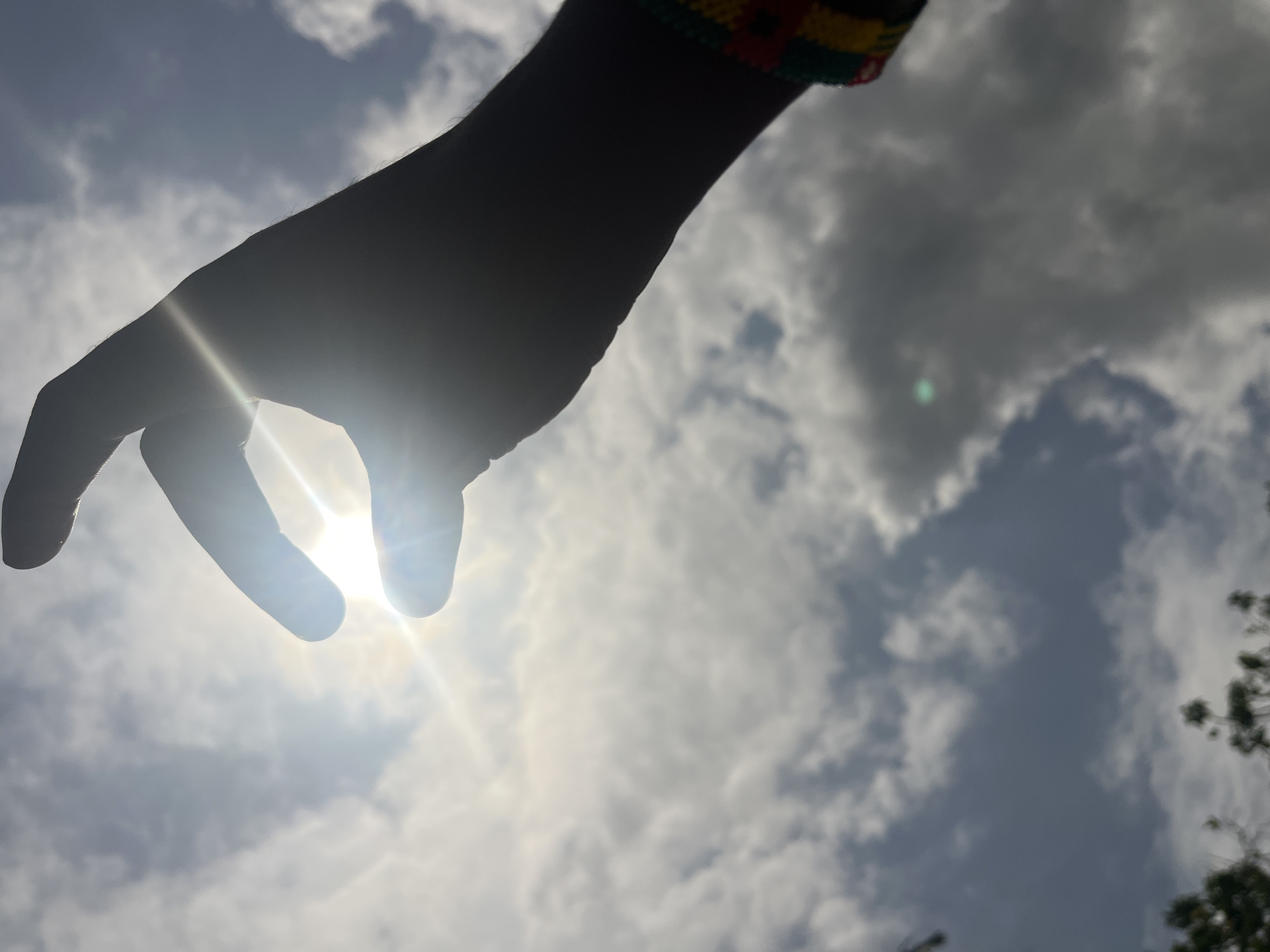 The image presents a striking low-angle shot of a human hand and forearm silhouetted against a bright, cloudy sky. The hand is positioned centrally, reaching downwards with fingers slightly curled, appearing to grasp or interact with the intense sun. A powerful lens flare emanates from the sun, which is centrally positioned within the outline of the hand, creating a dazzling burst of light.
The strong backlighting renders the hand and arm almost completely black, highlighting their form against the luminous sky. A notable detail is a colorful wristband or part of a sleeve visible on the upper right arm, featuring distinct red, yellow, and green bands, possibly with additional patterns.
The background shows a sky with varied cloud cover, including both bright white and darker grey clouds, indicating a partially overcast but still sunny day. The intensity of the sun suggests it is daytime. A small amount of dark tree foliage is visible on the far right edge of the frame, hinting at an outdoor natural setting. The photograph emphasizes the dramatic contrast between light and shadow and captures the brilliance of the sun framed by the human hand. No visible text is present in the image. The location context for this media is Ikot Ekpene, Nigeria.