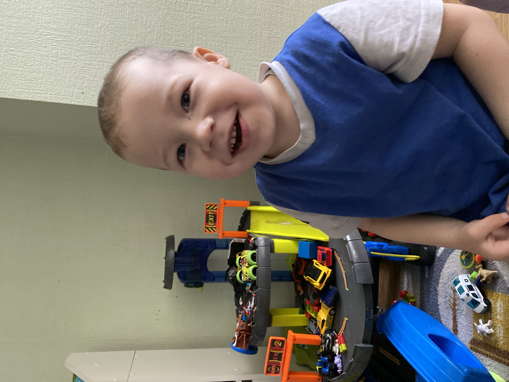 A young child with short, light hair and wearing a blue and white shirt is smiling and looking at the camera. The child is positioned slightly above and to the right of a multi-level toy car garage filled with toy cars. Various toy vehicles and animals are scattered on a patterned carpet near the garage. The background consists of a plain, light-colored wall. There is no indication of this being in Sochi, Russia.