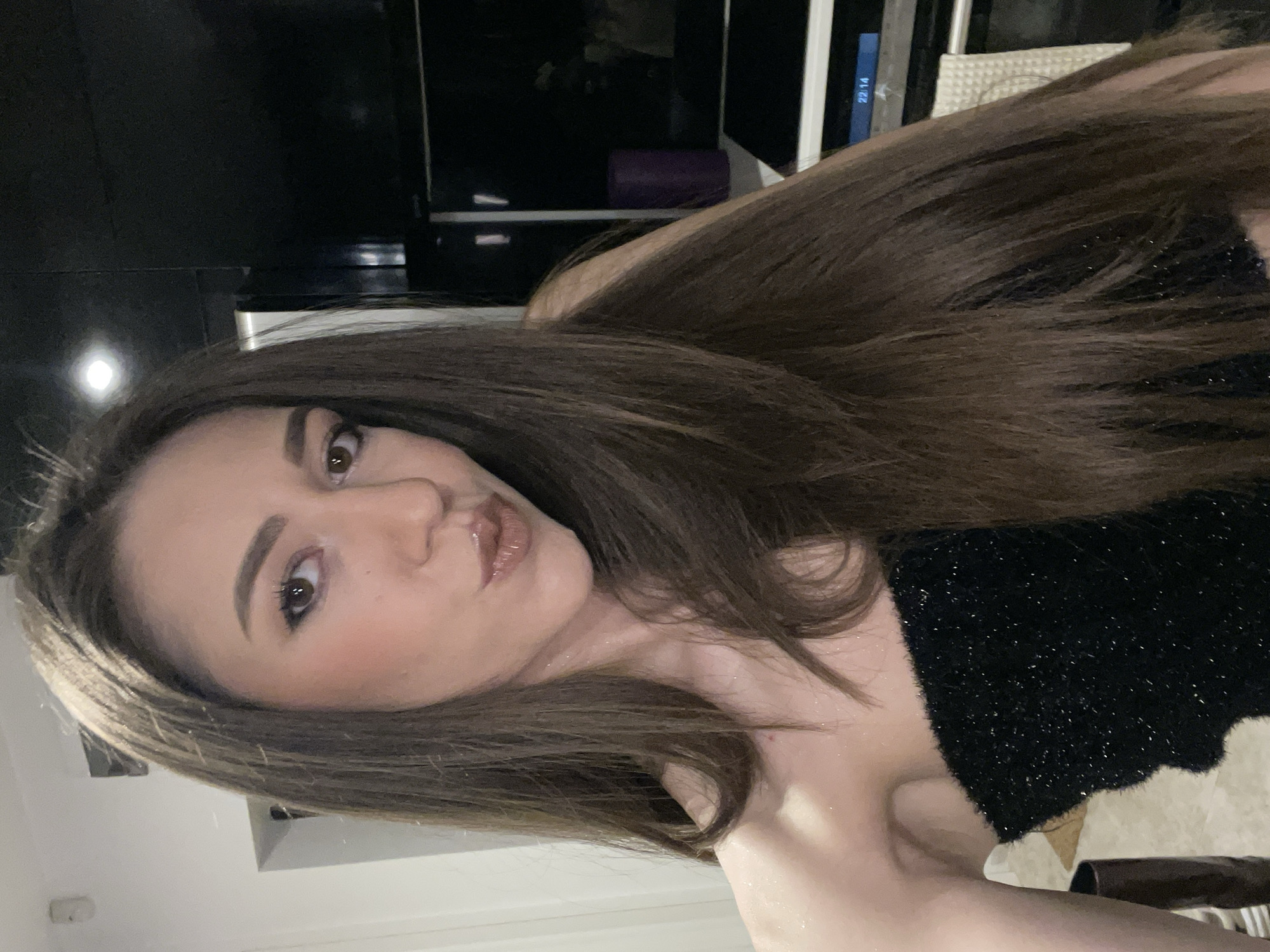 A young woman with long brown hair is posing for a selfie. She is wearing a black, sparkly top. Her makeup is visible and includes lip gloss. The background features a dark television screen and white walls. The location cannot be determined as Sofia, Bulgaria, from the visual information alone.