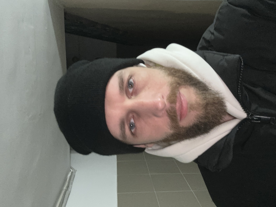 An adult male with light blue eyes and a reddish-brown beard is depicted in a headshot, facing the camera directly with a neutral expression. He wears a black knit beanie, a white hooded sweatshirt, and a black padded jacket, with a white earbud visible in his right ear. The background on the left features a tiled wall composed of light-colored rectangular tiles arranged horizontally. The background on the right is dark and indistinct. A metallic pipe or conduit runs horizontally along the ceiling-wall junction above the subject's left shoulder. The image captures an indoor utilitarian environment, possibly a hallway or stairwell, located in Saint Petersburg, Russia.