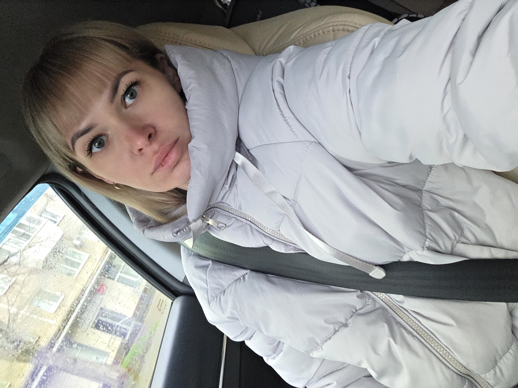 A young woman with blonde hair, bangs, and light eyes is the central subject of this image, captured inside a car in Chaikovsky, Russia. She is wearing a light grey or lavender puffer jacket, which appears partially zipped, and a dark seatbelt is visible across her chest. The woman is positioned diagonally in the frame, looking directly towards the camera with a calm, neutral expression. Behind her, a light-colored car seat or headrest is visible.
Through the car window on the left side of the frame, multi-story residential or commercial buildings and the bare branches of a tree can be seen, indicating an urban or suburban setting. The car window itself is covered with numerous raindrops and streaks, suggesting recent rain or ongoing precipitation, consistent with the warm puffer jacket. Natural light illuminates the scene, indicating it is daytime. Faint, blurry text, likely a manufacturer's label or identification numbers (possibly "PILKINGTON"), is barely discernible on the car window.